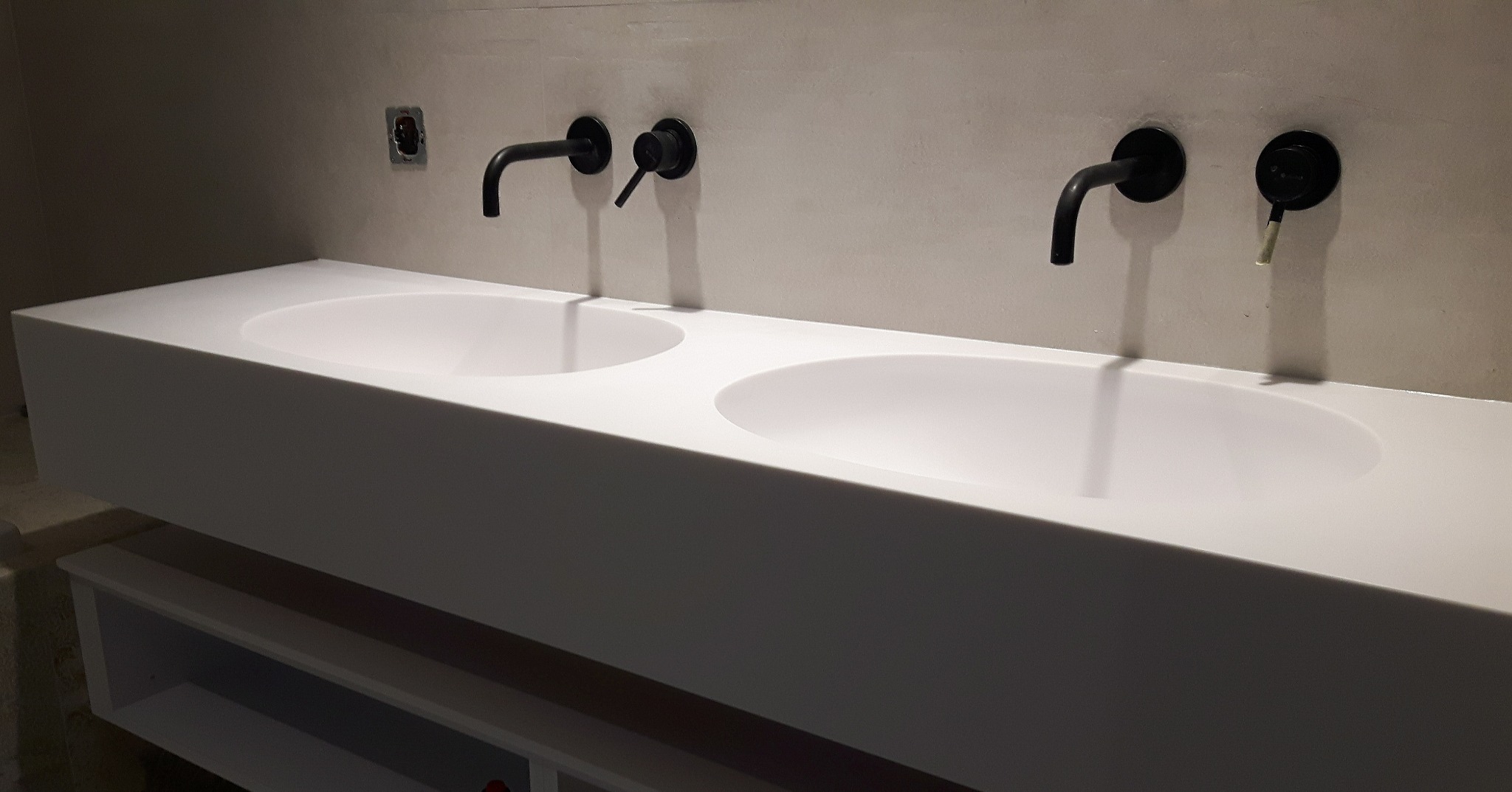 Podwójna, biała umywalka z czarnymi, ściennymi bateriami w nowoczesnej łazience z surowym wykończeniem ścian.