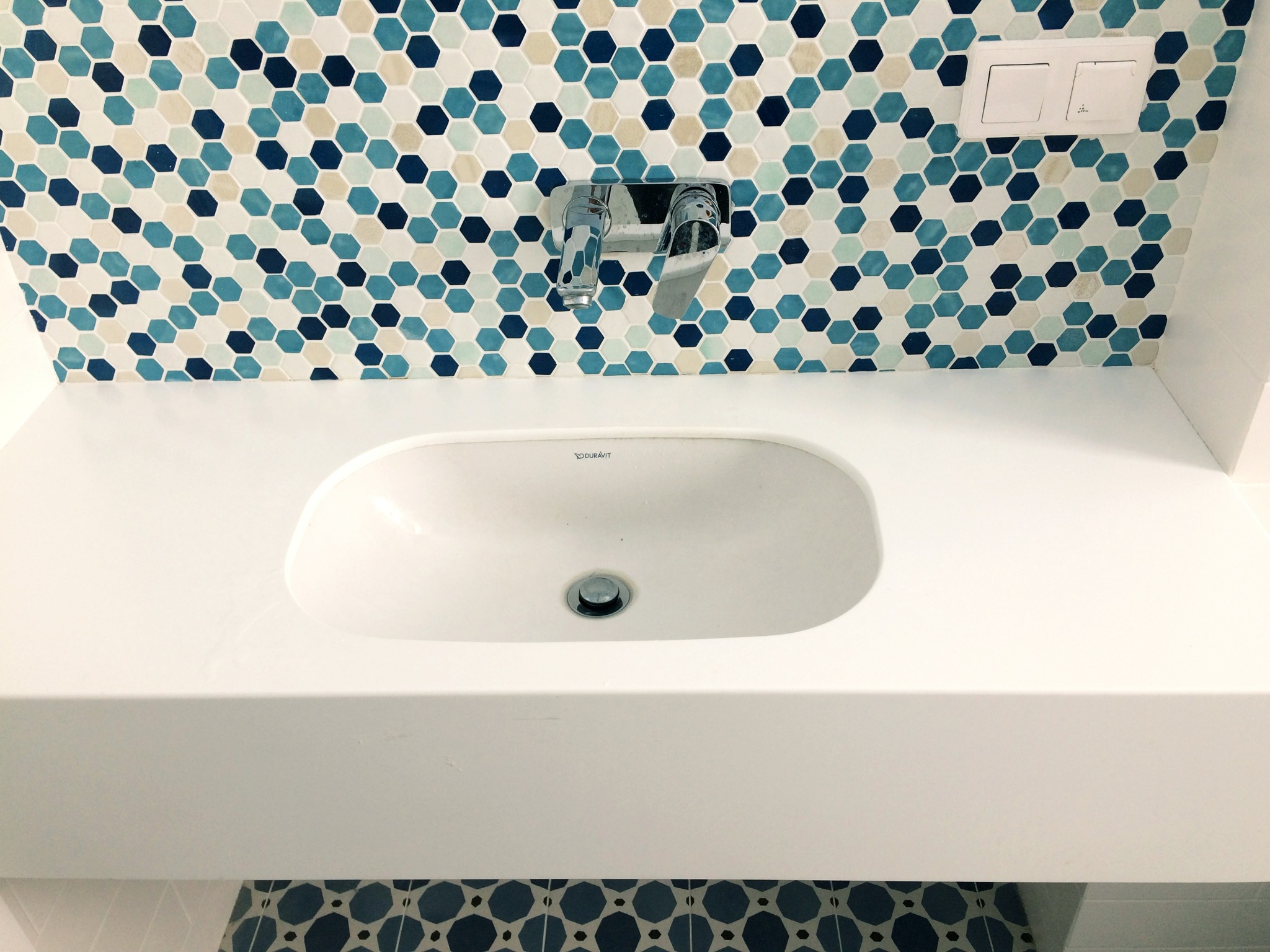 Biała, prostokątna umywalka z chromowaną baterią na tle mozaiki z heksagonów w odcieniach błękitu i bieli, z widocznym fragmentem toalety z identyczną mozaiką na podłodze.