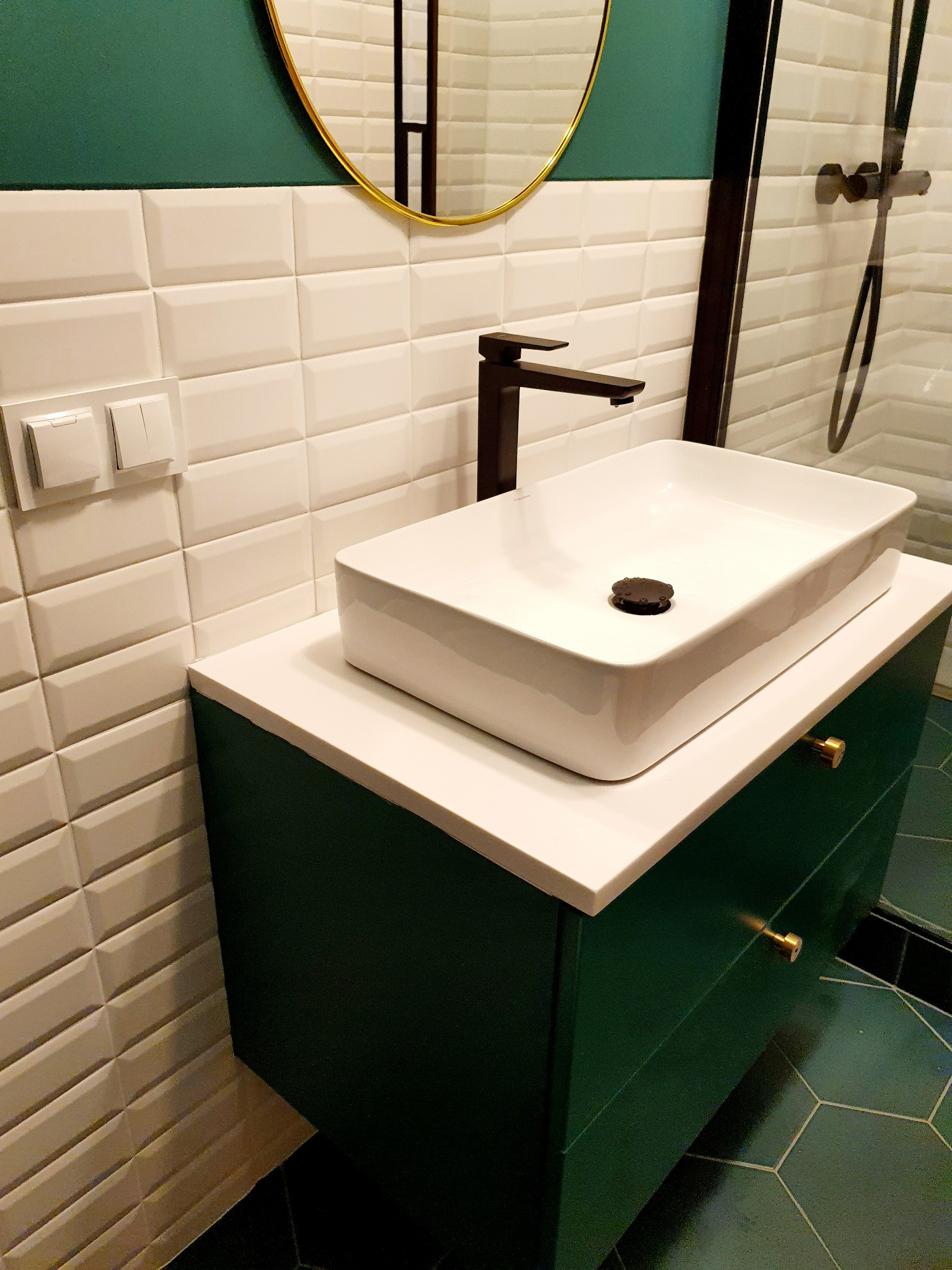 Nowoczesna łazienka z białą, prostokątną umywalką nablatową, czarną armaturą i zieloną szafką z złotymi uchwytami na tle białych, trójwymiarowych płytek ściennych i heksagonalnych płytek podłogowych.