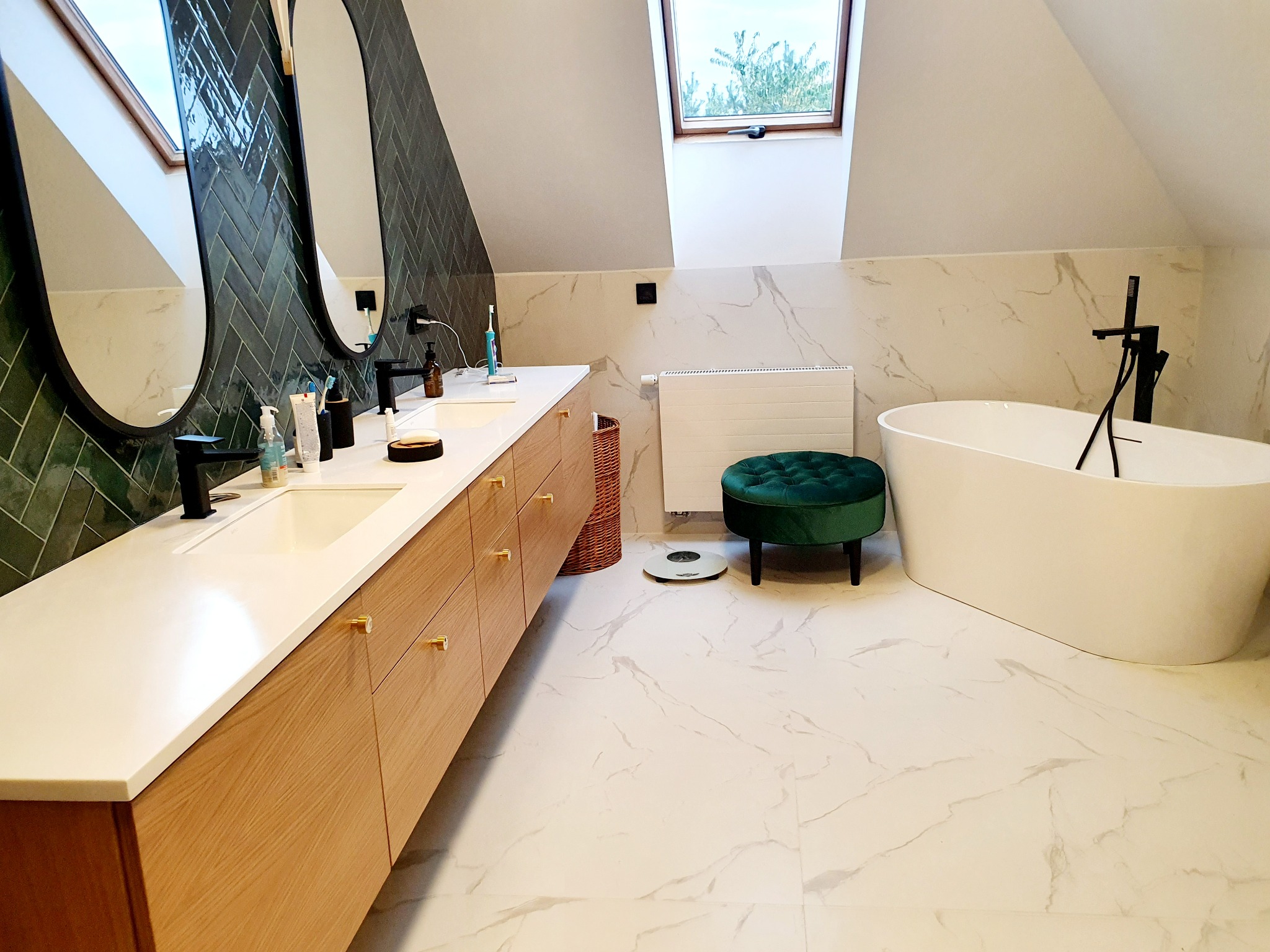 Nowoczesna łazienka na poddaszu z długim drewnianym blatem, owalnymi lustrami, zieloną mozaiką na ścianie i wolnostojącą białą wanną.