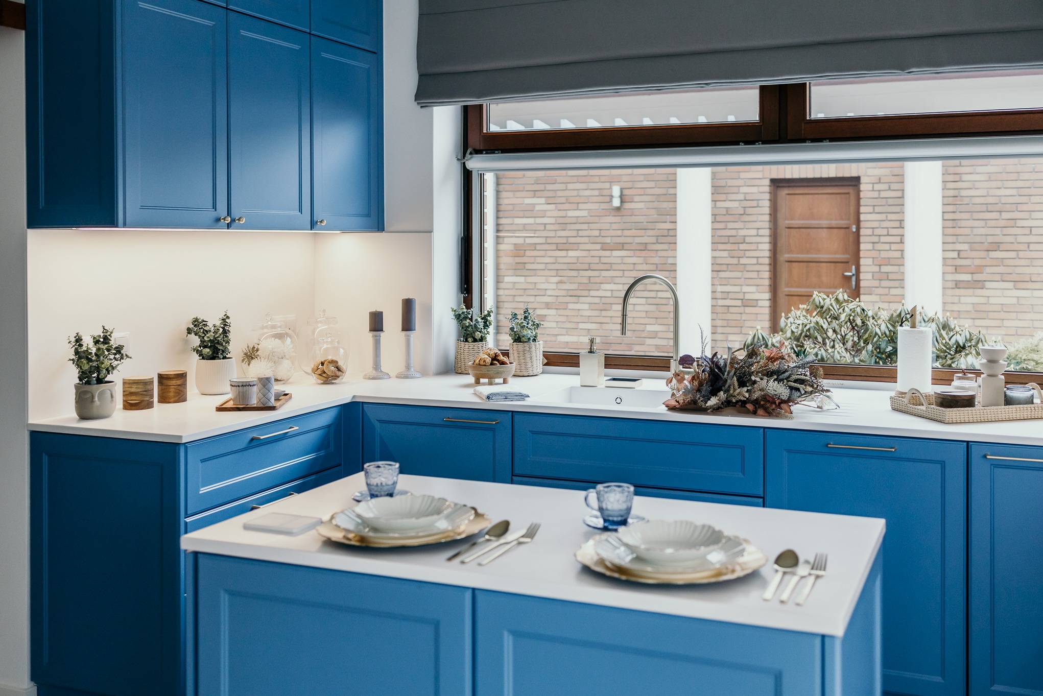 Niebieska kuchnia z białym blatem, nakryty stół na wyspie kuchennej, widok na ceglany budynek przez okno.