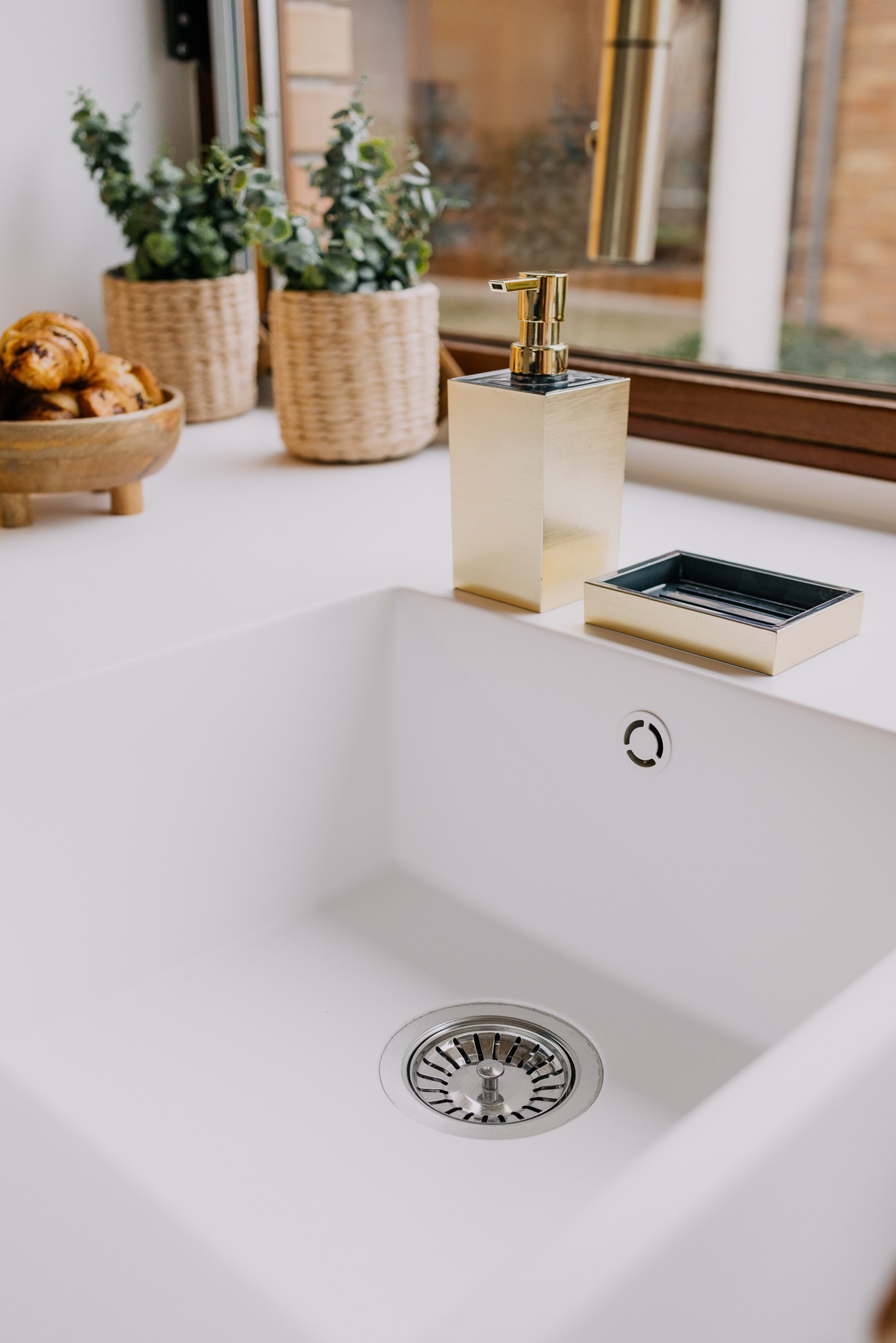 Biały zlew kuchenny z ociekaczem, złotym dozownikiem mydła i mydelniczką, na blacie ciastka i rośliny w plecionych doniczkach, widok z góry.