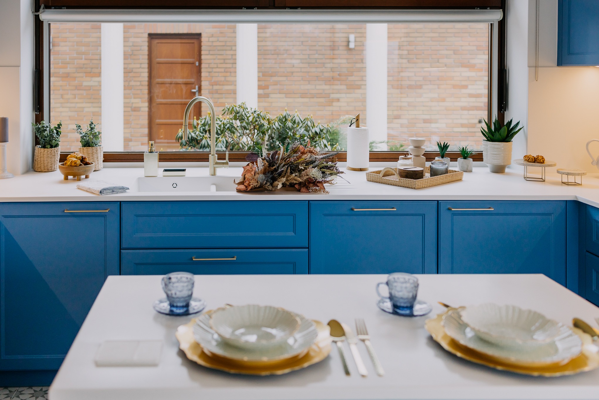 Biały blat kuchenny z niebieskimi szafkami, dekoracjami i zastawą stołową w słonecznym wnętrzu.