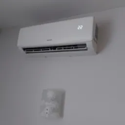 Biała klimatyzacja Sinclair zamontowana na jasnej ścianie, wyświetlająca temperaturę 22 stopnie. Pod nią kinkiet z motywem kwiatowym.