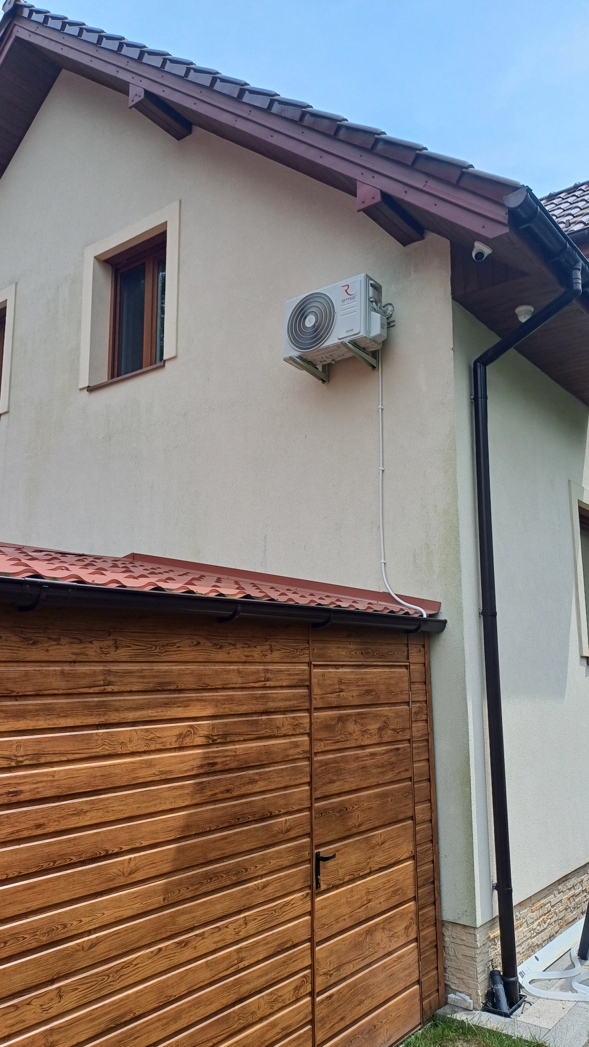 Zewnętrzna jednostka klimatyzacji zamontowana na elewacji domu, widoczna instalacja odprowadzająca skropliny, brązowa brama garażowa z poziomych paneli w tle.