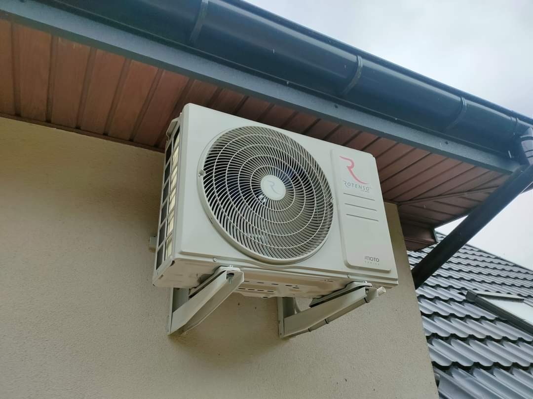 Zewnętrzna jednostka klimatyzacji Rotenso zamontowana na ścianie budynku pod okapem z widocznymi elementami mocującymi.