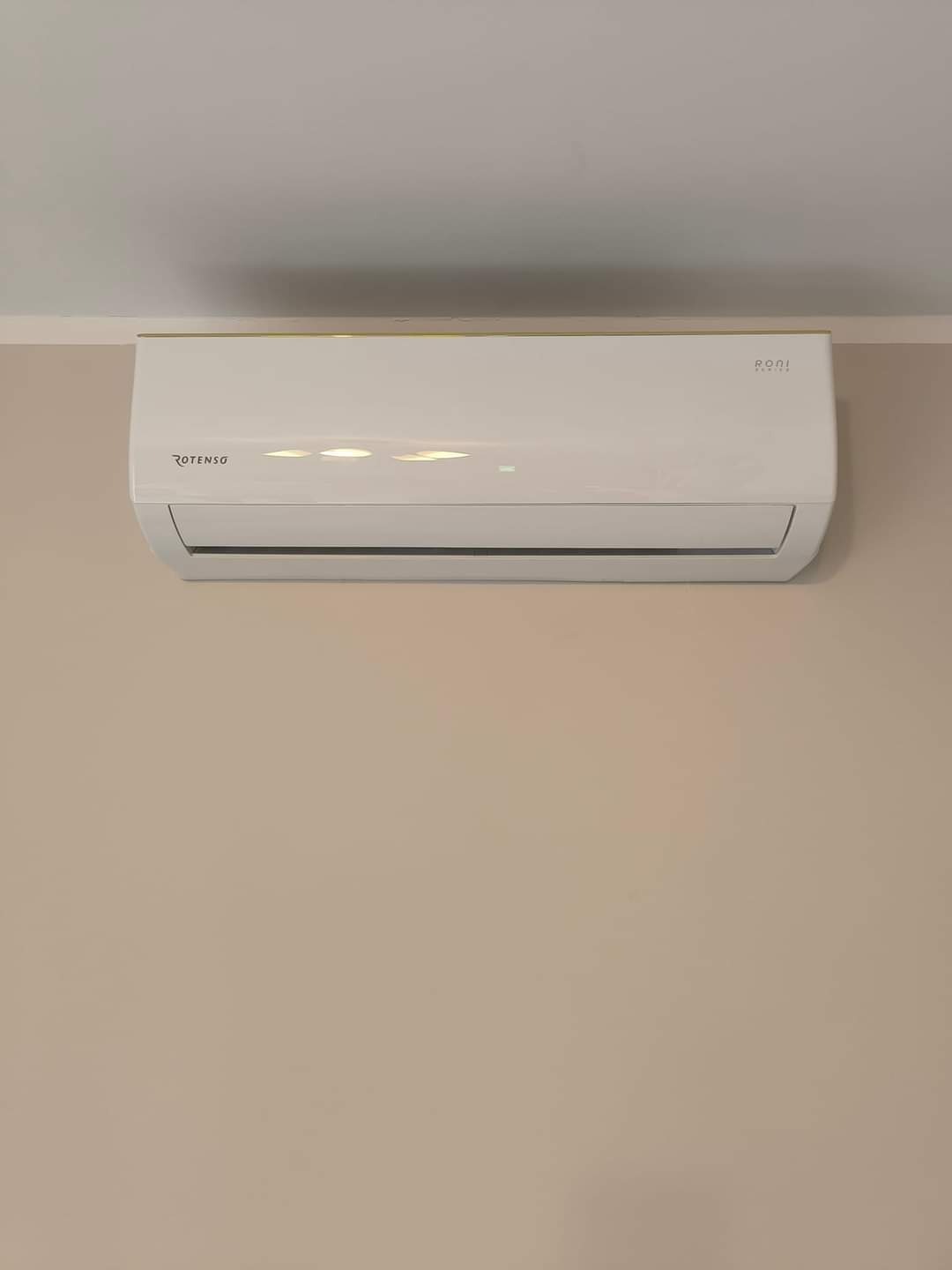 Biały klimatyzator ROTENSO Roni Series zamontowany na ścianie o beżowym odcieniu, widoczne diody sygnalizacyjne.