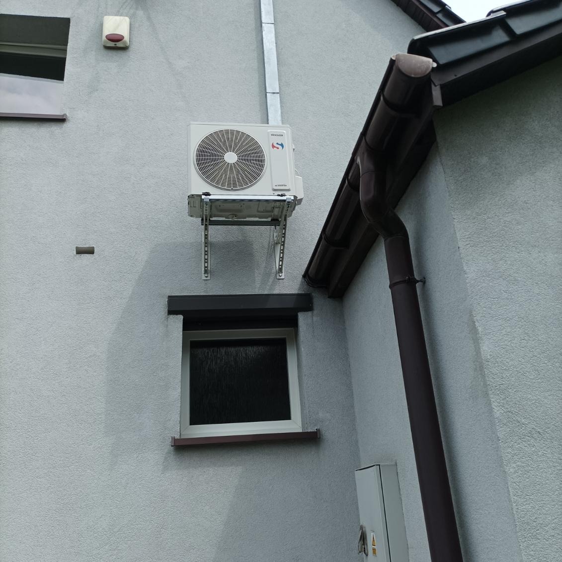 Zewnętrzna jednostka klimatyzacji zamontowana na wspornikach na elewacji domu, widoczna rynna i okno z roletą.