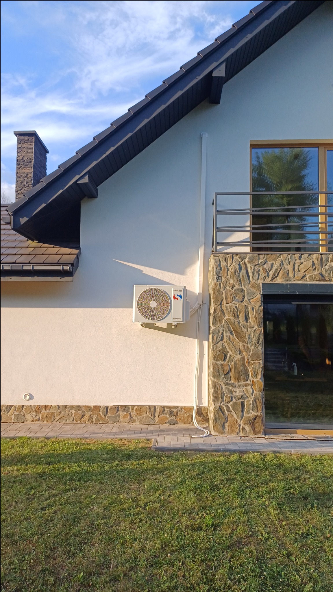 Zewnętrzna jednostka klimatyzatora zamontowana na elewacji domu z elementami kamiennymi, widoczne przewody instalacyjne i trawnik.