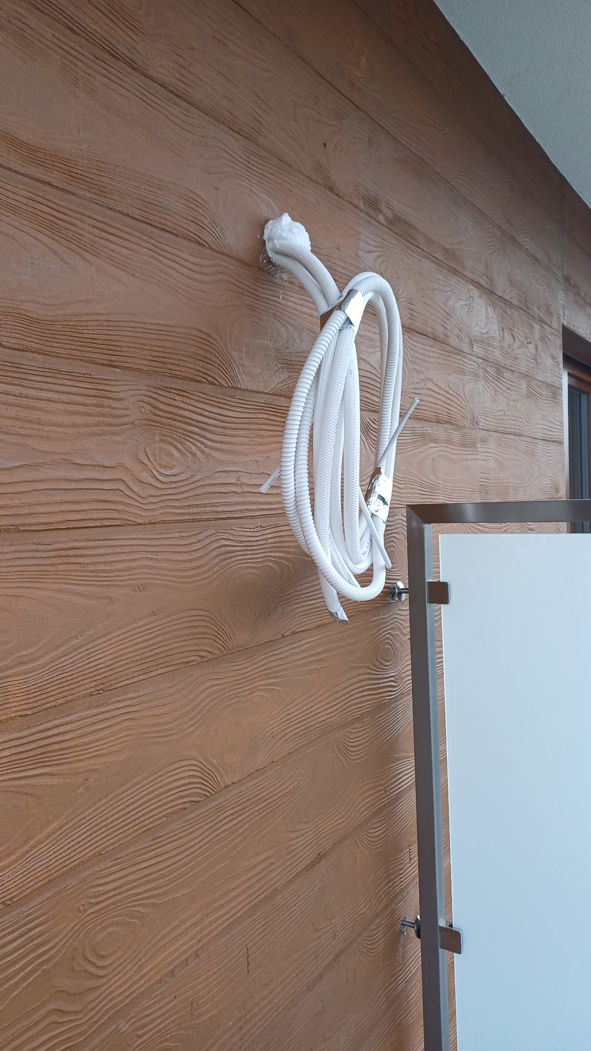 Białe rurki instalacyjne przymocowane do brązowej ściany imitującej drewno, zabezpieczone srebrną taśmą i pianką montażową.