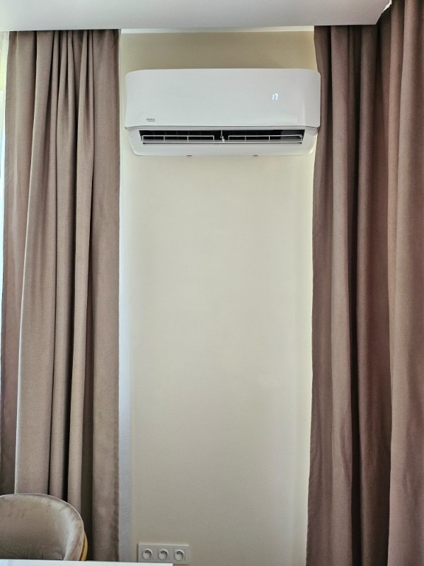 Biały klimatyzator ścienny z wyświetlaczem temperatury, zamontowany na kremowej ścianie, otoczony brązowymi zasłonami, widoczne gniazdka elektryczne pod urządzeniem.