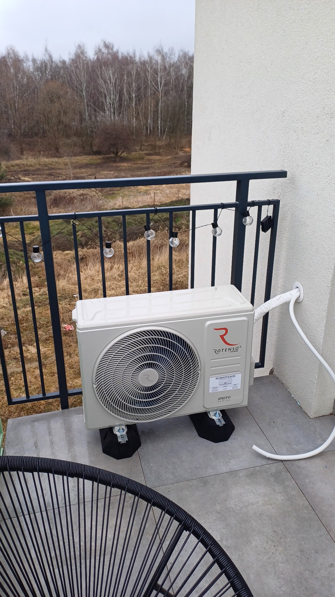 Jednostka zewnętrzna klimatyzacji Rotenso zamontowana na balkonie w Knurowie, widok na las za balustradą, dekoracyjne lampki wiszące na balustradzie.