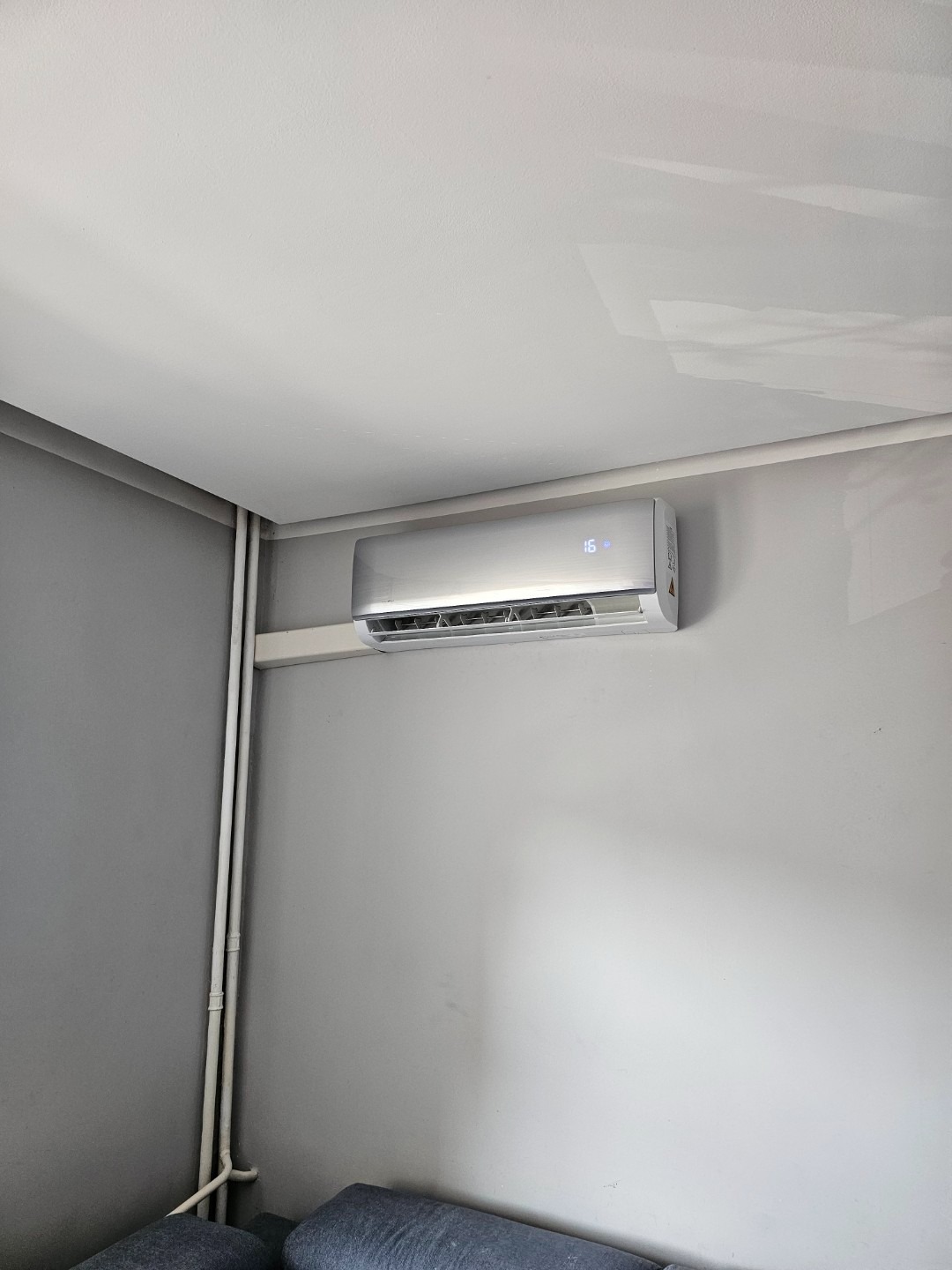 Klimatyzator ścienny zamontowany w rogu pokoju, widoczne rury instalacyjne, ustawiony na 16 stopni Celsjusza.
