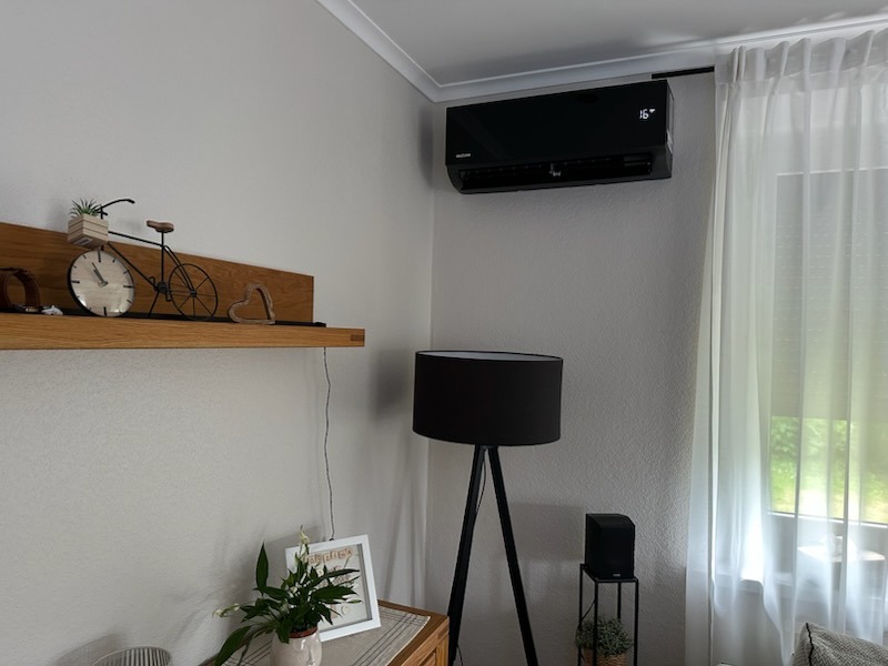 Czarny klimatyzator ścienny zamontowany w rogu pokoju, obok okna z białymi zasłonami i lampy na trójnogu. Na ścianie półka z dekoracjami.