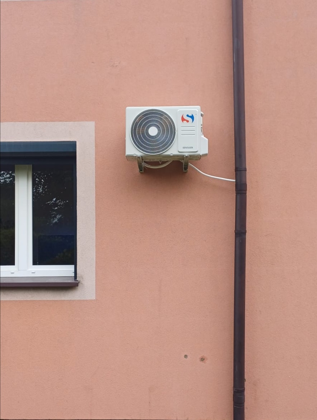 Biały klimatyzator na różowej ścianie budynku obok okna i rury spustowej. Widoczne mocowania urządzenia i kabel zasilający.
