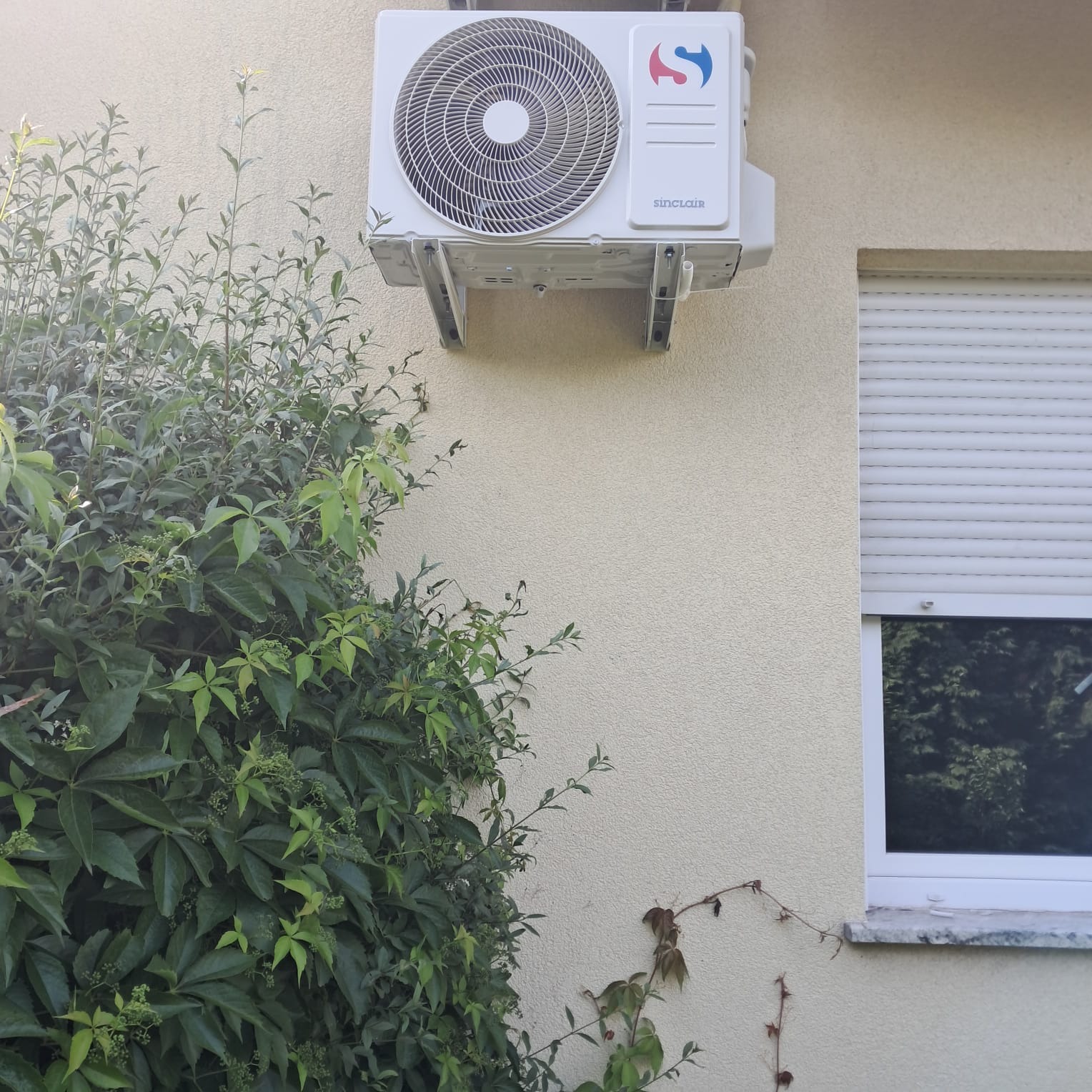 Zamontowany klimatyzator Sinclair na beżowej elewacji budynku, widoczny zza zieleni, obok okna z opuszczonymi roletami.