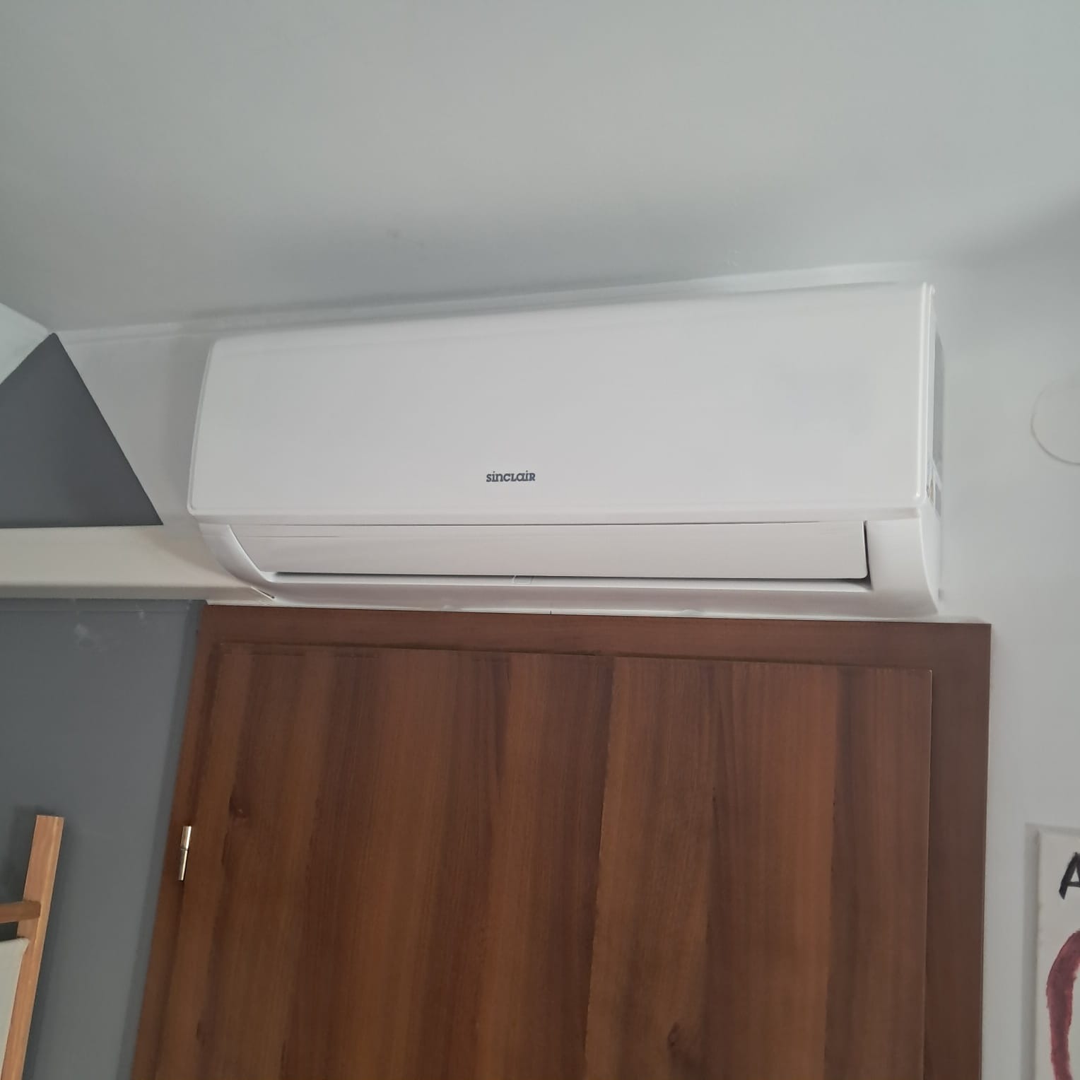 Biała klimatyzacja Sinclair, zamontowana nad drewnianą szafą, na tle szarej ściany z elementami dekoracyjnymi. Widoczny fragment drabiny.