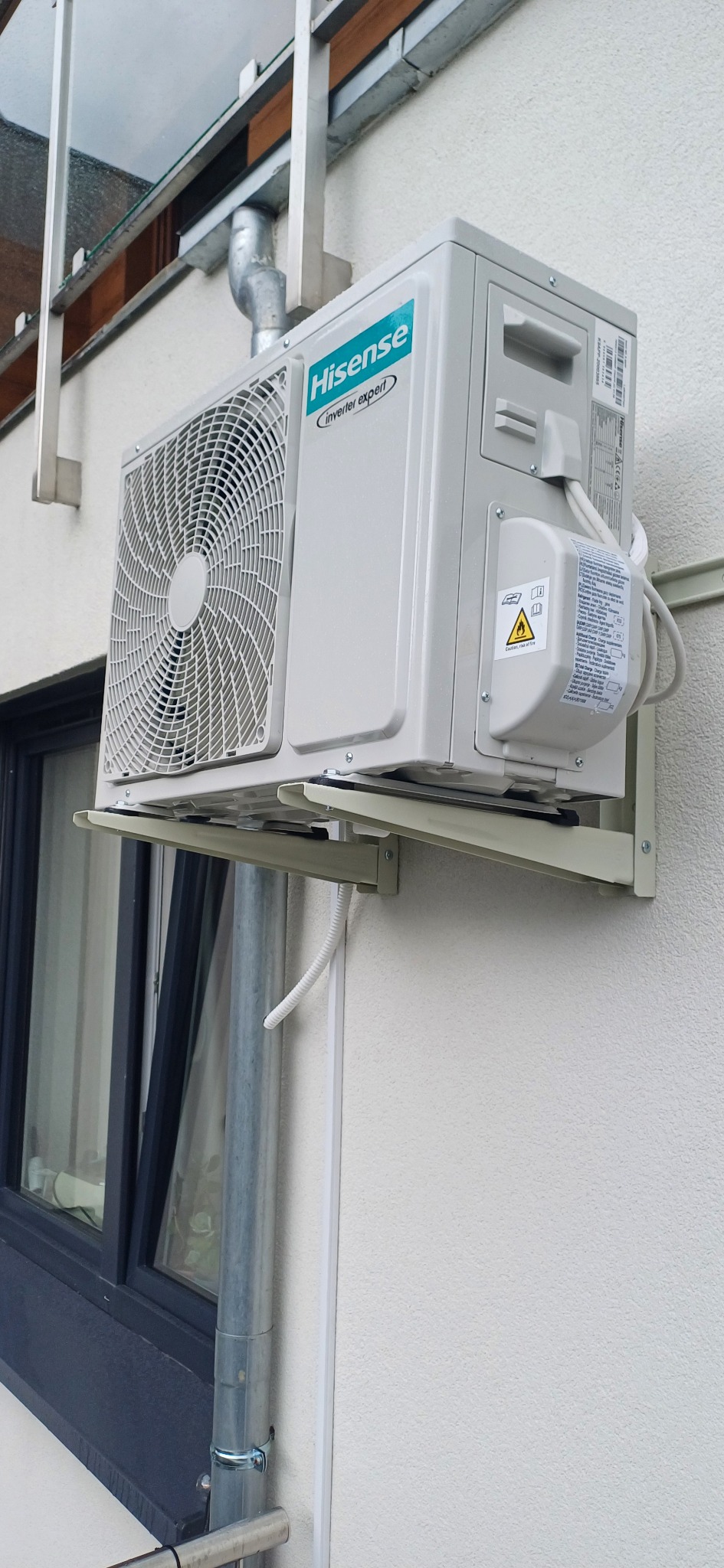 Zewnętrzna jednostka klimatyzacji Hisense Inverter Expert, zamontowana na wsporniku przy ścianie budynku, obok okna i rury spustowej.