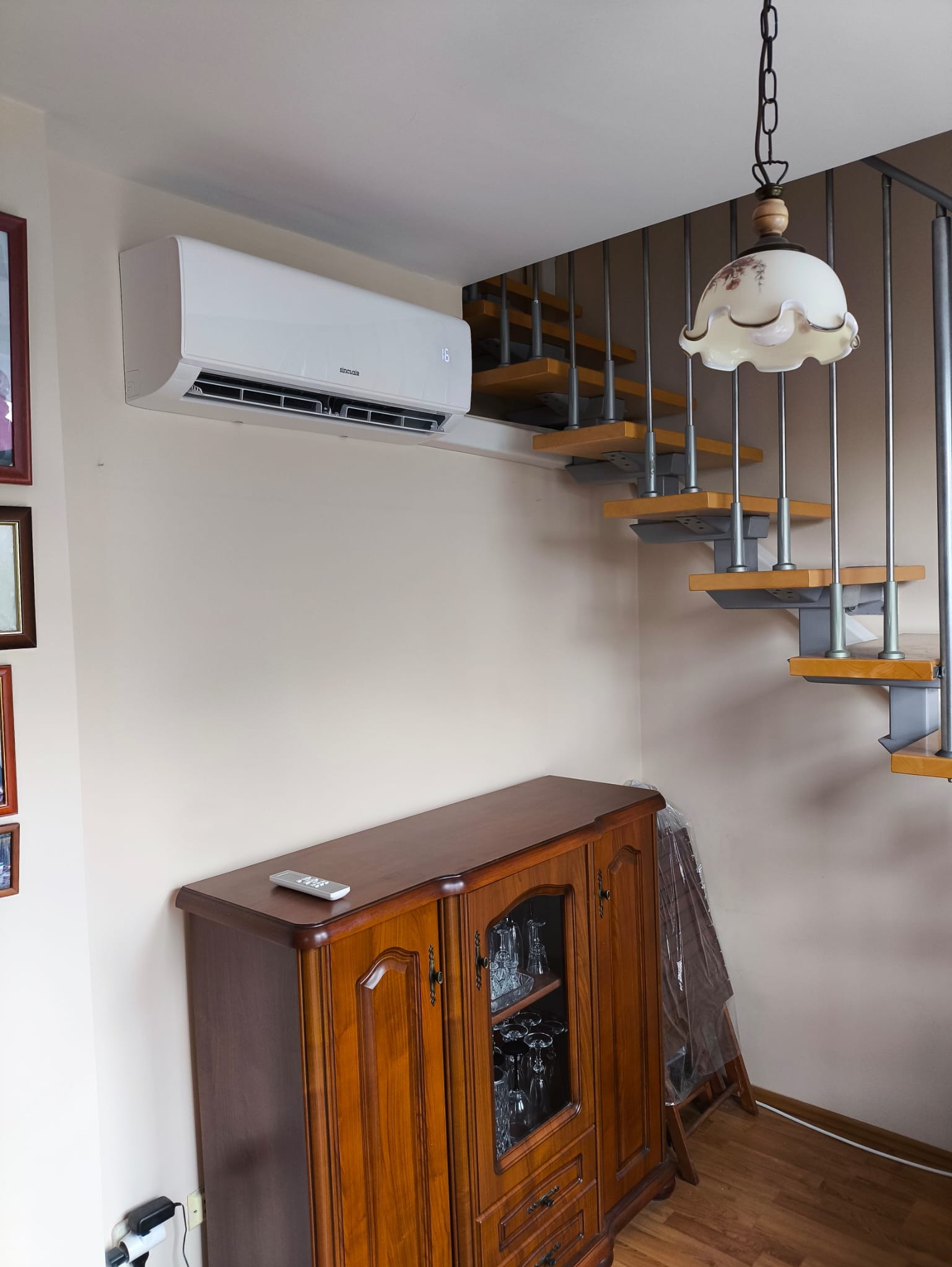 Biała klimatyzacja Sinclair na ścianie w pokoju z drewnianymi schodami, komodą i lampą. Pilot leży na komodzie. Widok na wnętrze domu.
