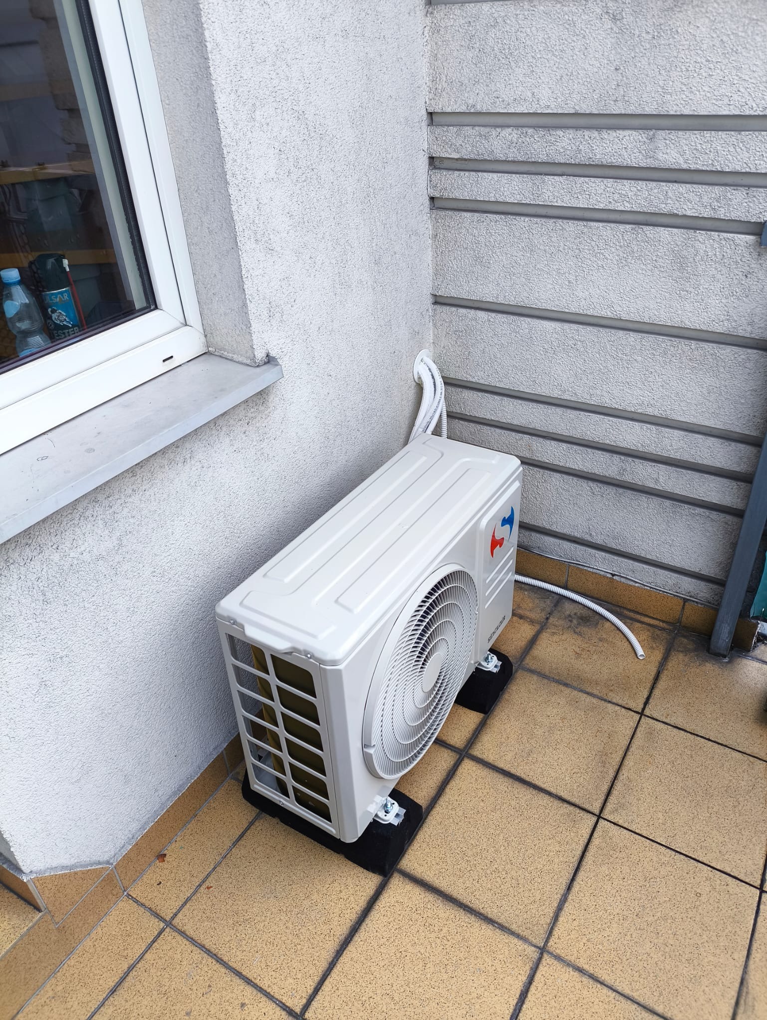 Biały klimatyzator zewnętrzny zamontowany na balkonie, widoczne przewody i wsporniki. Tło stanowi ściana z szarym tynkiem i fragment okna.