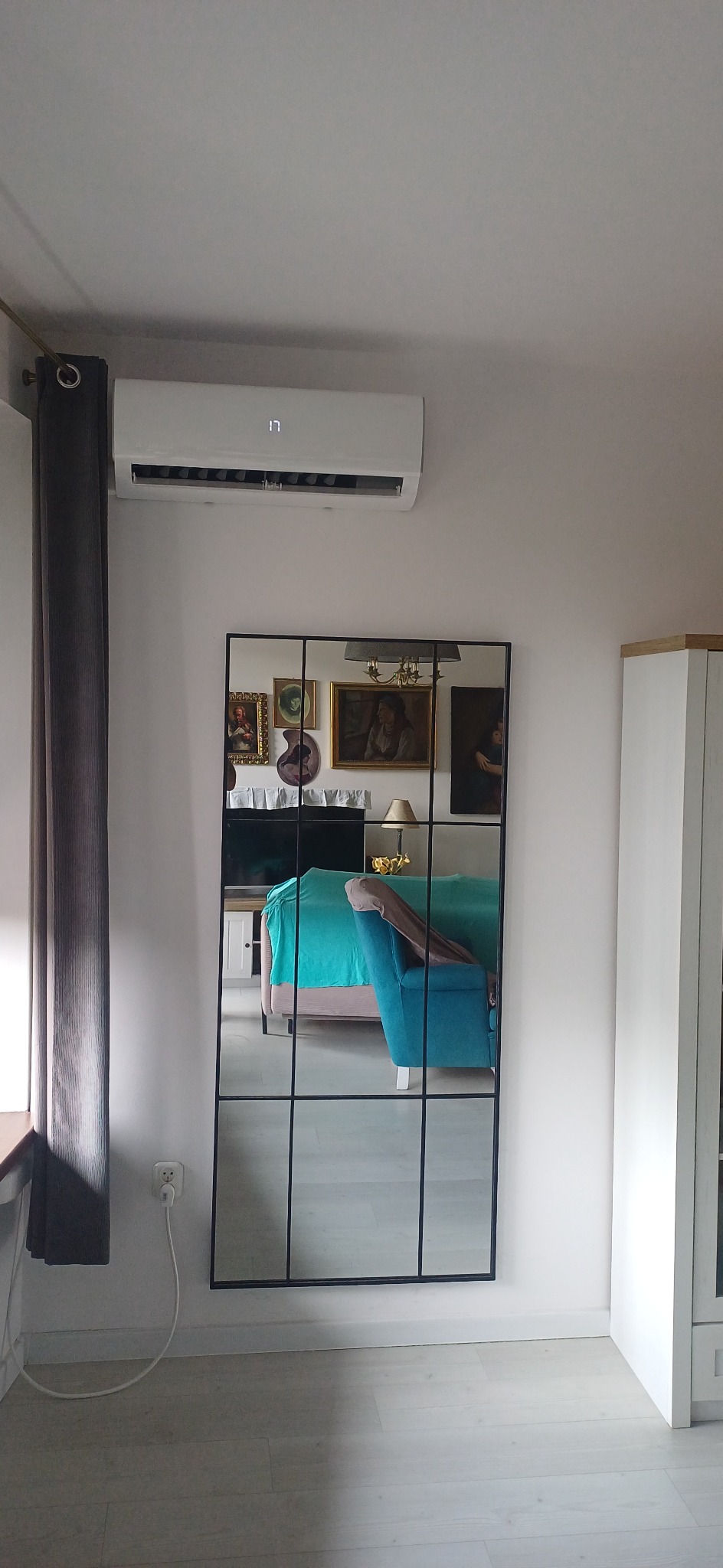 Biały klimatyzator ścienny nad lustrem w czarnej ramie, odbijającym turkusowy fotel i obrazy w salonie. Widoczny fragment zasłony i mebla.