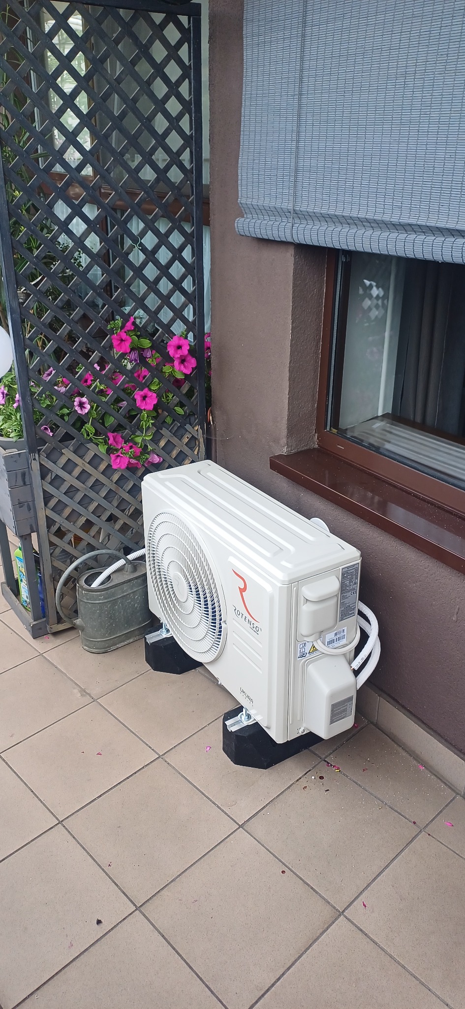 Biała jednostka zewnętrzna klimatyzacji Rotenso UKURA, zamontowana na czarnych podkładkach na tarasie. Obok kratka z kwiatami i zwinięta roleta okienna.