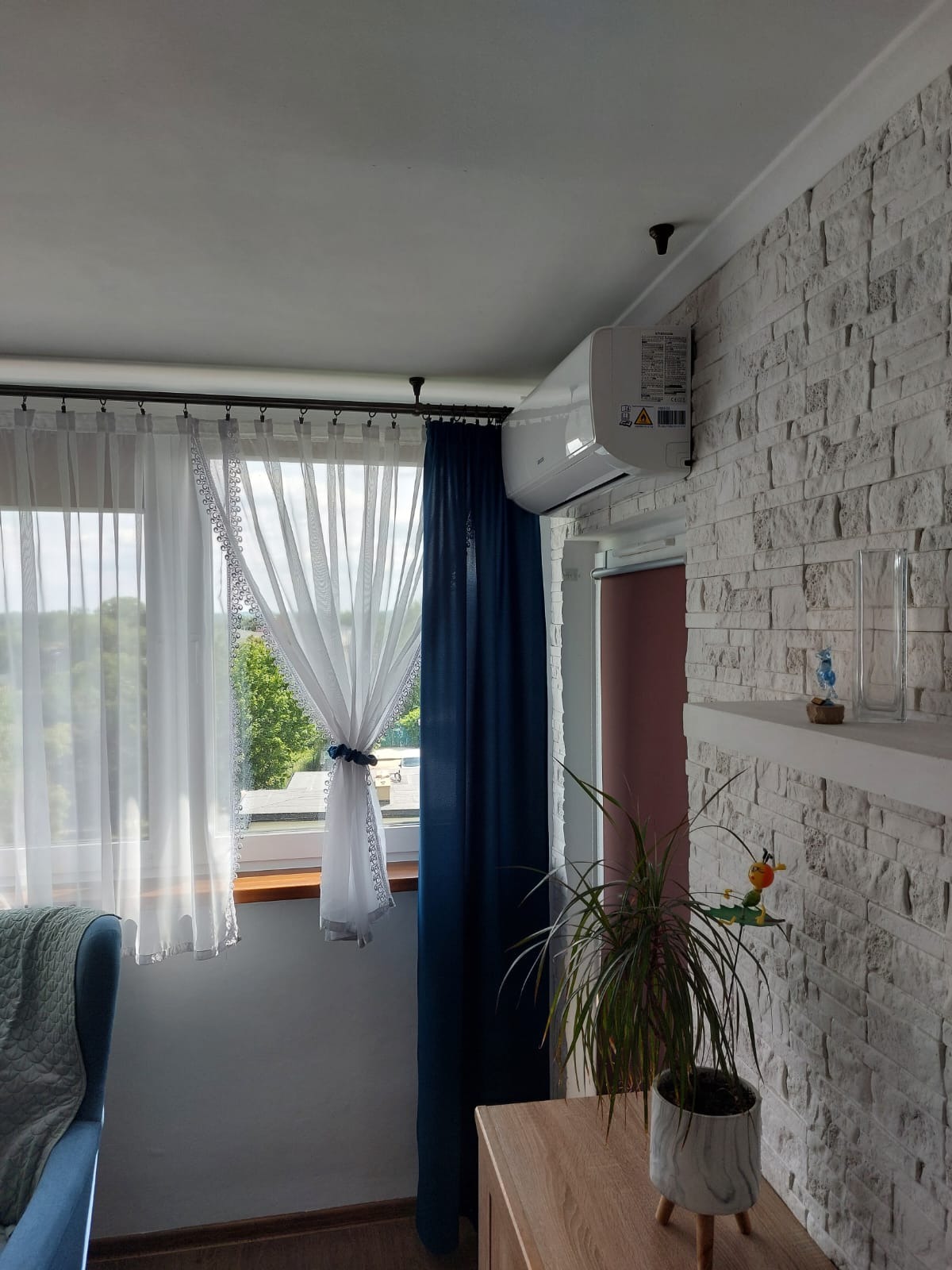 Biała klimatyzacja na ścianie z dekoracyjnej cegły w jasnym pokoju z zasłonami. Widok na zewnątrz przez okno. Wnętrze dopełnia roślina doniczkowa.