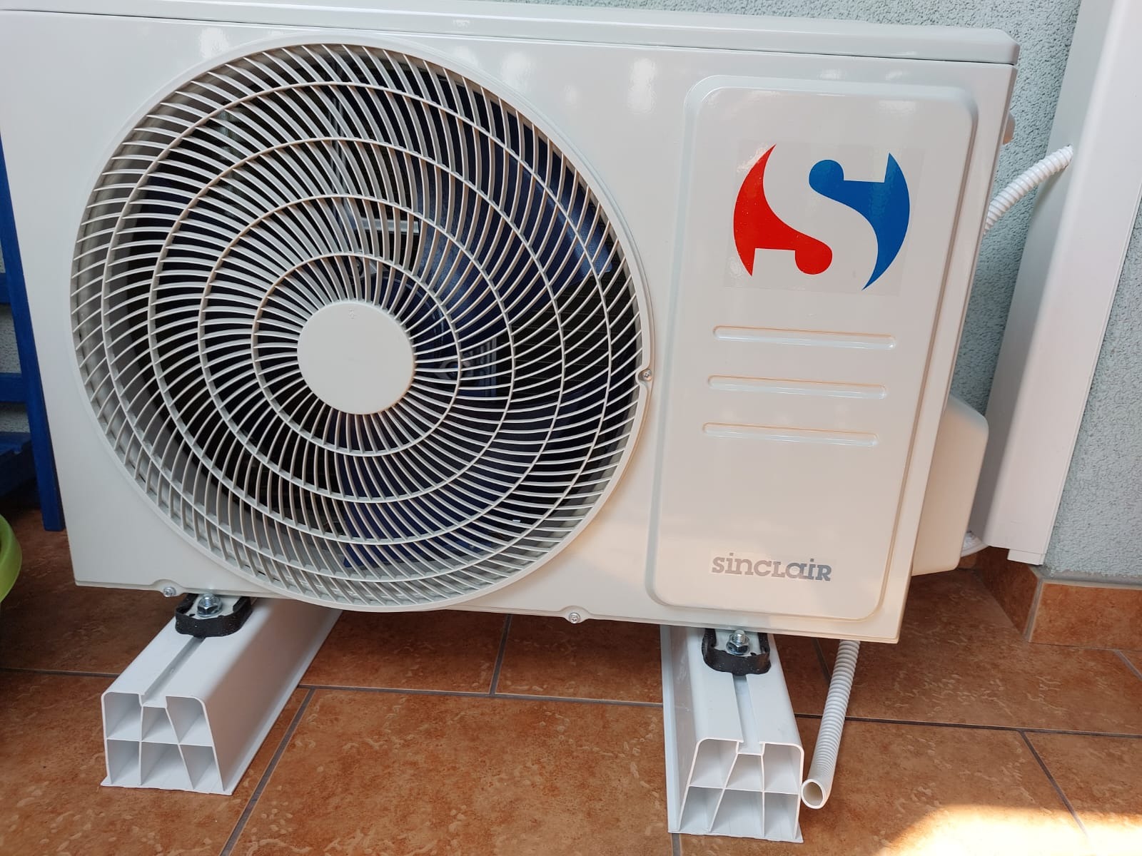 Biała jednostka zewnętrzna klimatyzacji Sinclair zamontowana na wspornikach z tworzywa, widoczna na tle brązowych płytek podłogowych i fragmentu szarej ściany.