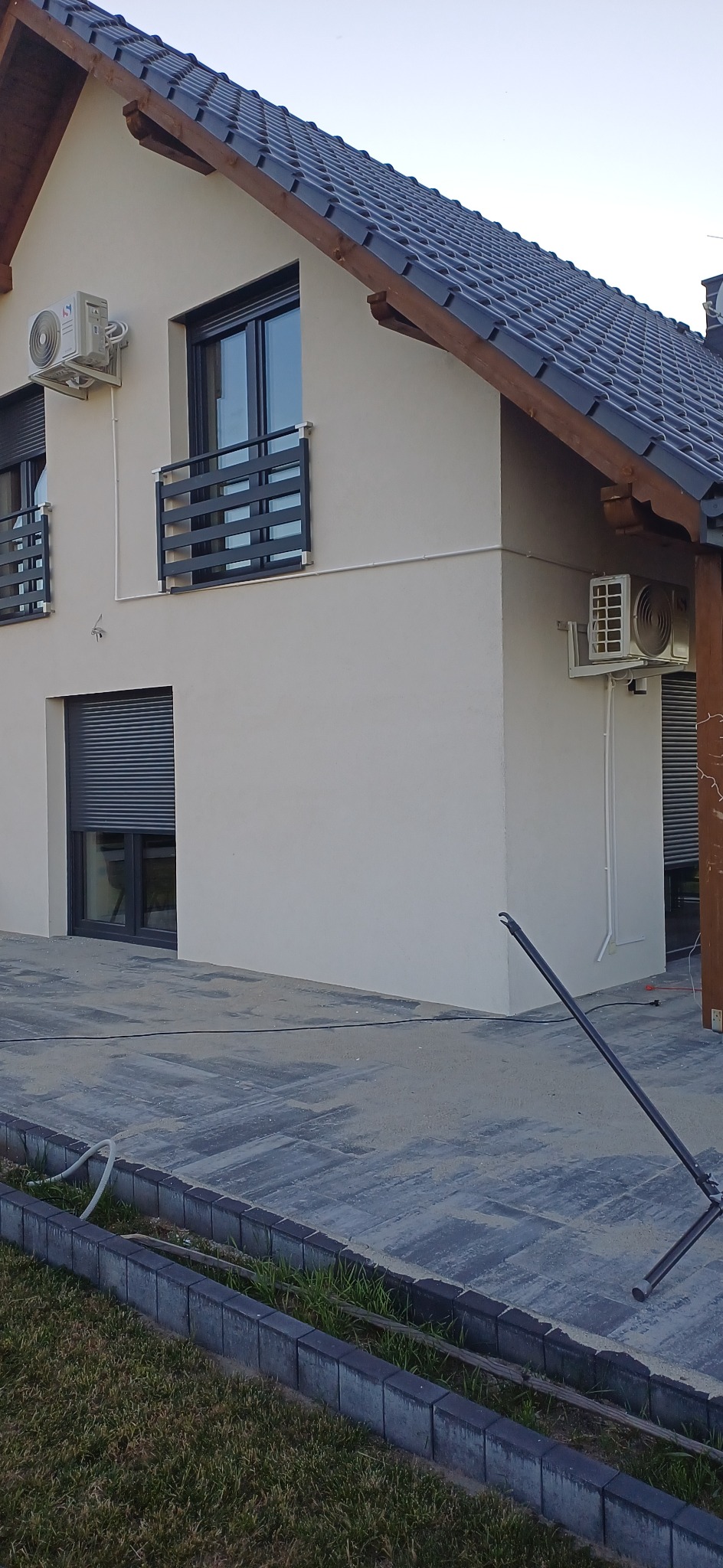 Elewacja domu z dwoma zainstalowanymi klimatyzatorami, widok z zewnątrz. Na pierwszym planie kostka brukowa i trawnik. Okna z roletami zewnętrznymi.