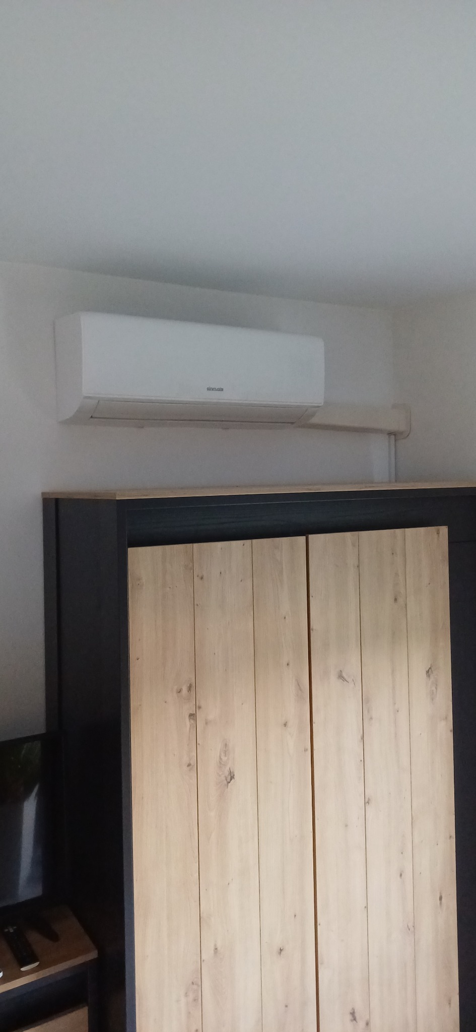 Biała jednostka klimatyzacyjna zamontowana na ścianie nad szafą z drewnianymi drzwiami w jasnym pomieszczeniu. Widoczne przewody instalacyjne.