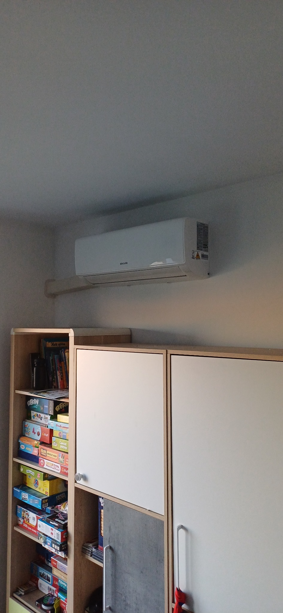 Biała klimatyzacja ścienna zamontowana nad regałem z grami planszowymi w pokoju. Widoczne detale montażu i etykiety ostrzegawcze na urządzeniu.