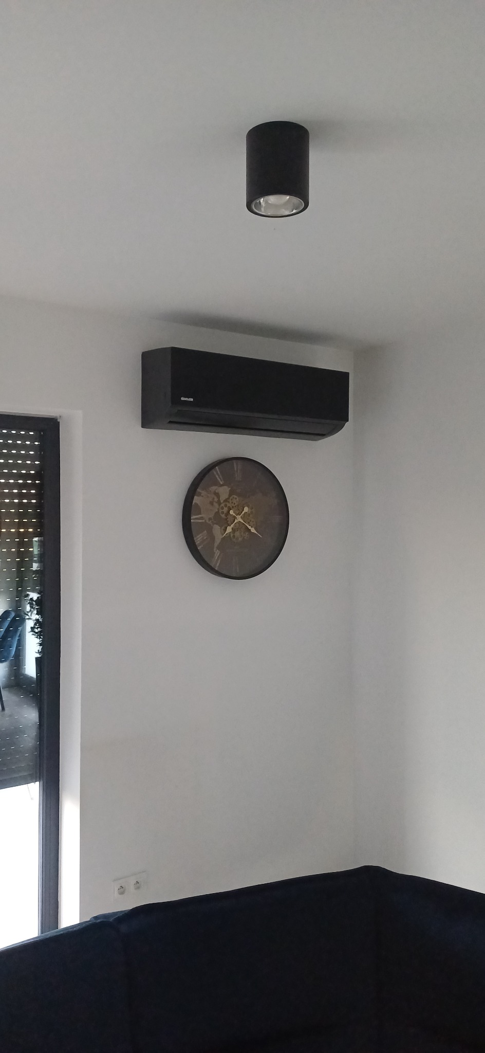 Czarna klimatyzacja ścienna w nowoczesnym wnętrzu, nad nią lampa sufitowa, obok okno z żaluzjami i dekoracyjny zegar. Minimalistyczny design.