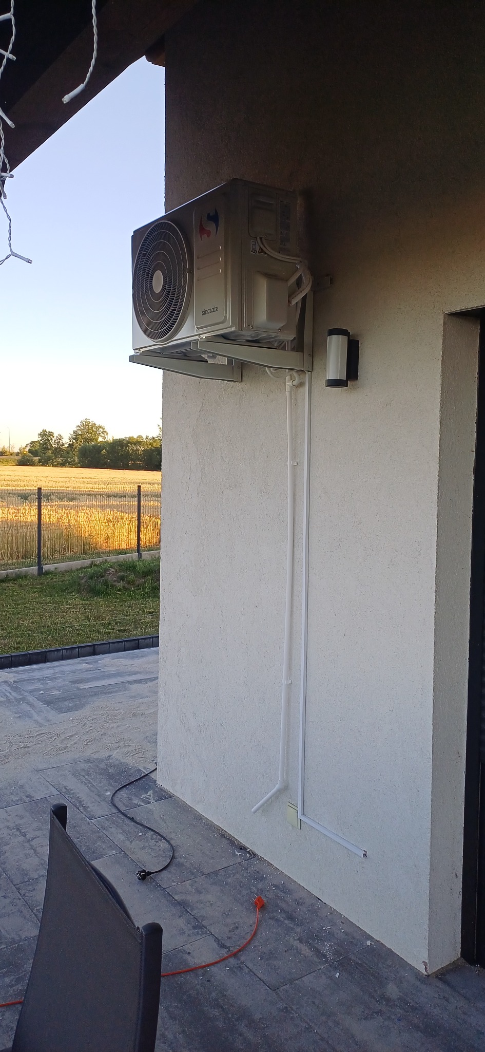 Zamontowany klimatyzator Sinclair na elewacji domu, z widocznymi rurami odprowadzającymi i lampą zewnętrzną. Tło stanowi pole i ogrodzenie.