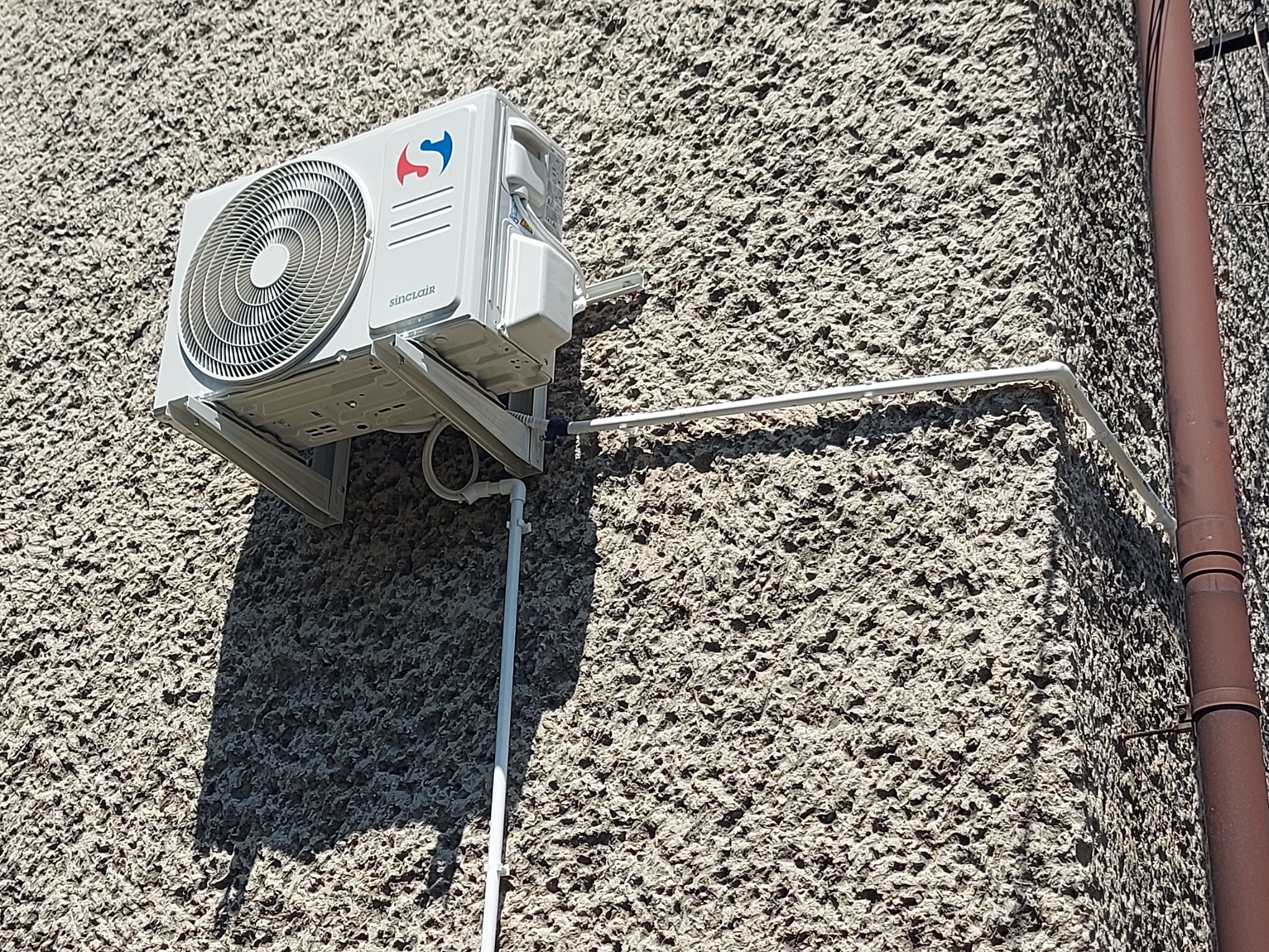 Biała jednostka zewnętrzna klimatyzacji Sinclair zamontowana na szorstkiej, jasnej elewacji budynku, z widocznymi rurami odprowadzającymi skropliny.