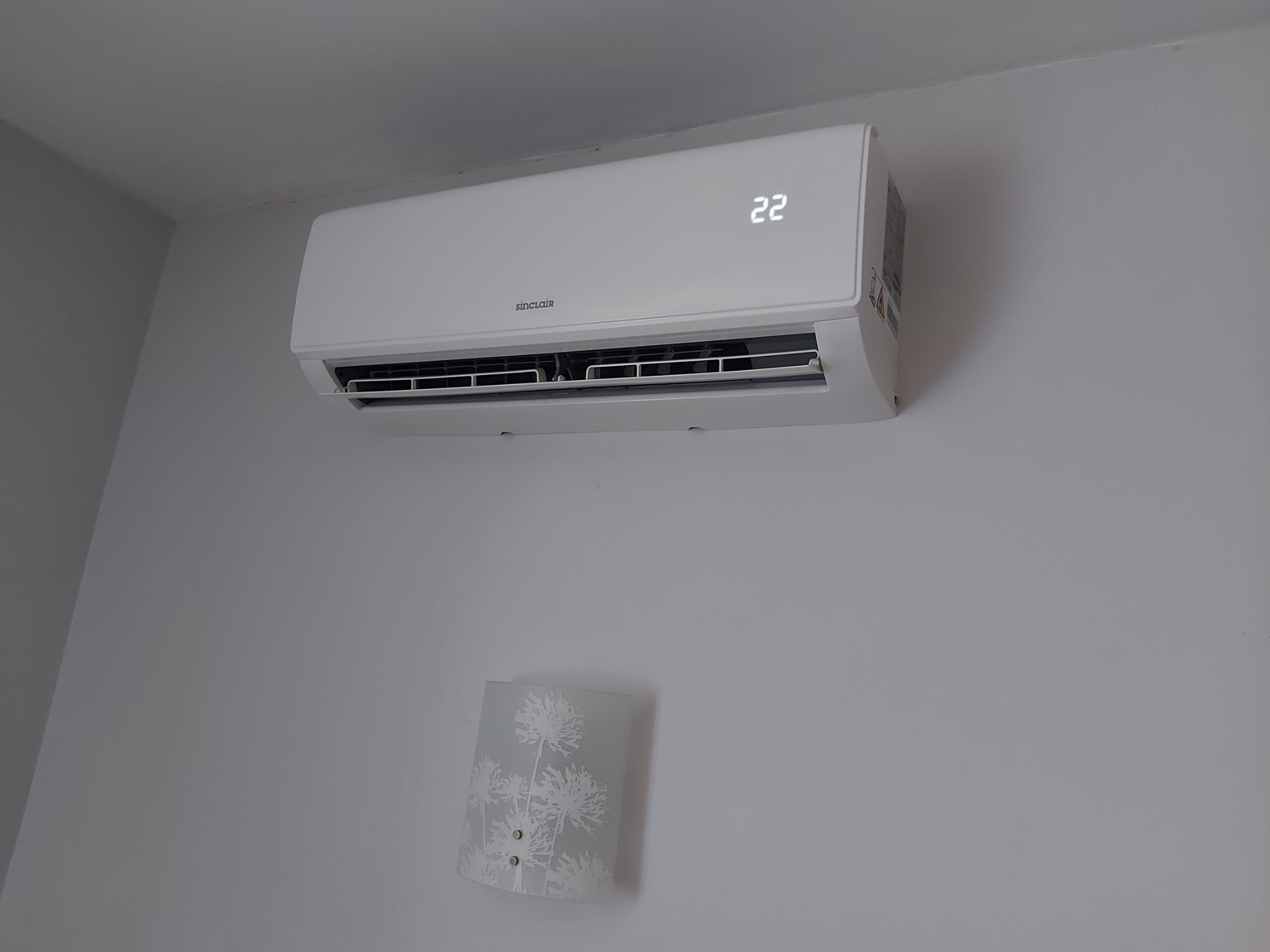 Biała klimatyzacja Sinclair zamontowana na jasnej ścianie, wyświetlająca temperaturę 22 stopnie. Pod nią kinkiet z motywem kwiatowym.
