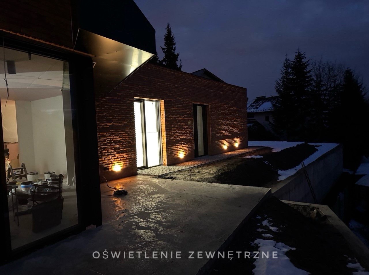 Nowoczesny dom z cegły z oświetleniem zewnętrznym LED wbudowanym w chodnik, widoczne okno z włączonym światłem oraz częściowo ośnieżony teren.