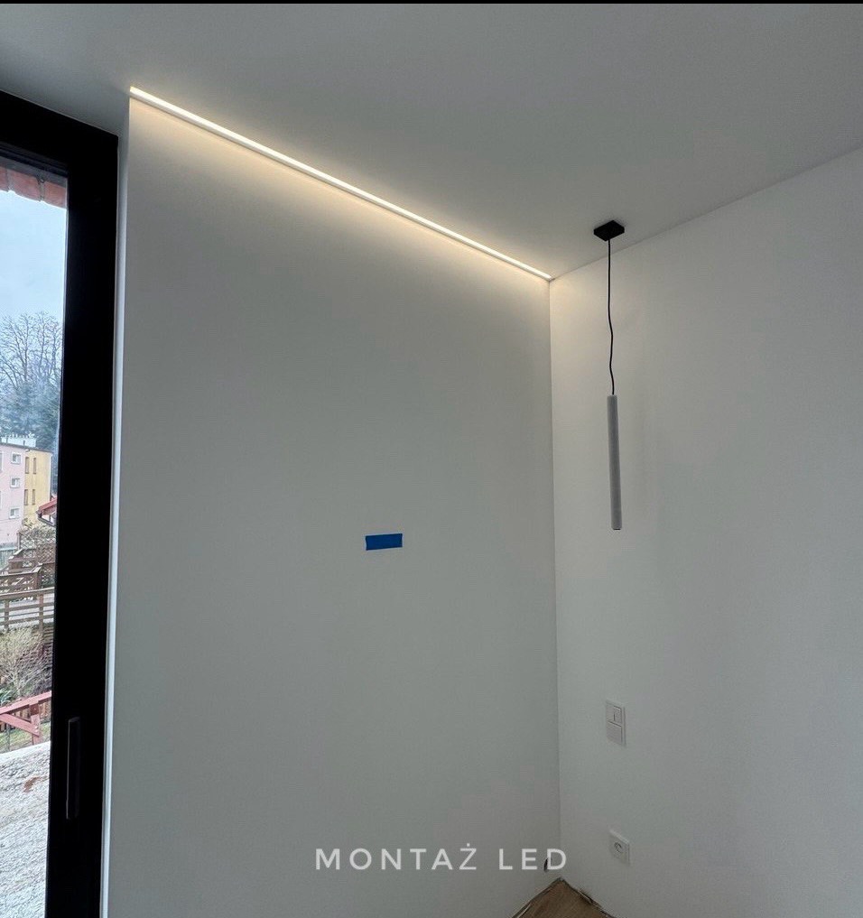 Nowoczesne oświetlenie LED wpuszczone w sufit, minimalistyczna lampa wisząca, surowy wystrój wnętrza z widocznym niebieskim fragmentem taśmy malarskiej na ścianie.