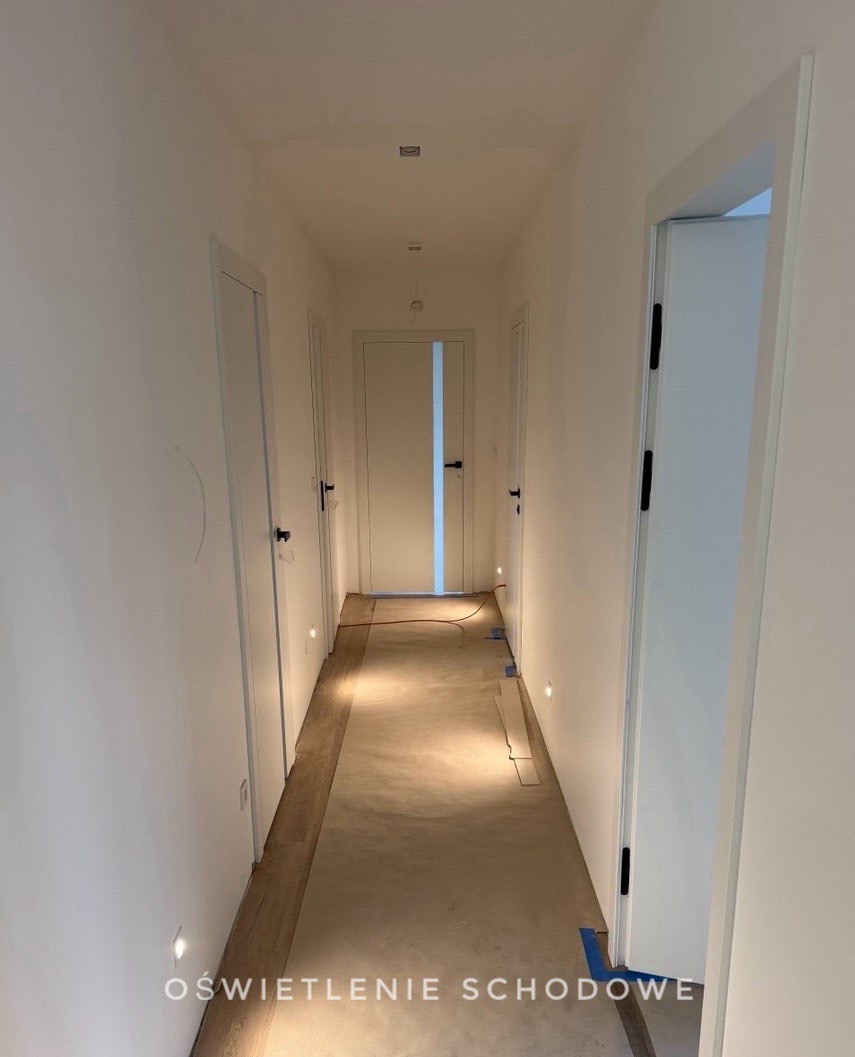 Długi korytarz z białymi ścianami i kilkoma parami drzwi. Na podłodze widoczne listwy i kable. Zamontowane oświetlenie punktowe przy podłodze i na suficie.