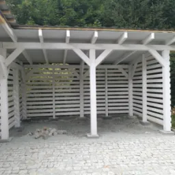 Biała, drewniana wiata z poziomymi listwami i kamienną podłogą z kostki granitowej, otoczona zielenią. Elementy konstrukcyjne w jasnym odcieniu.