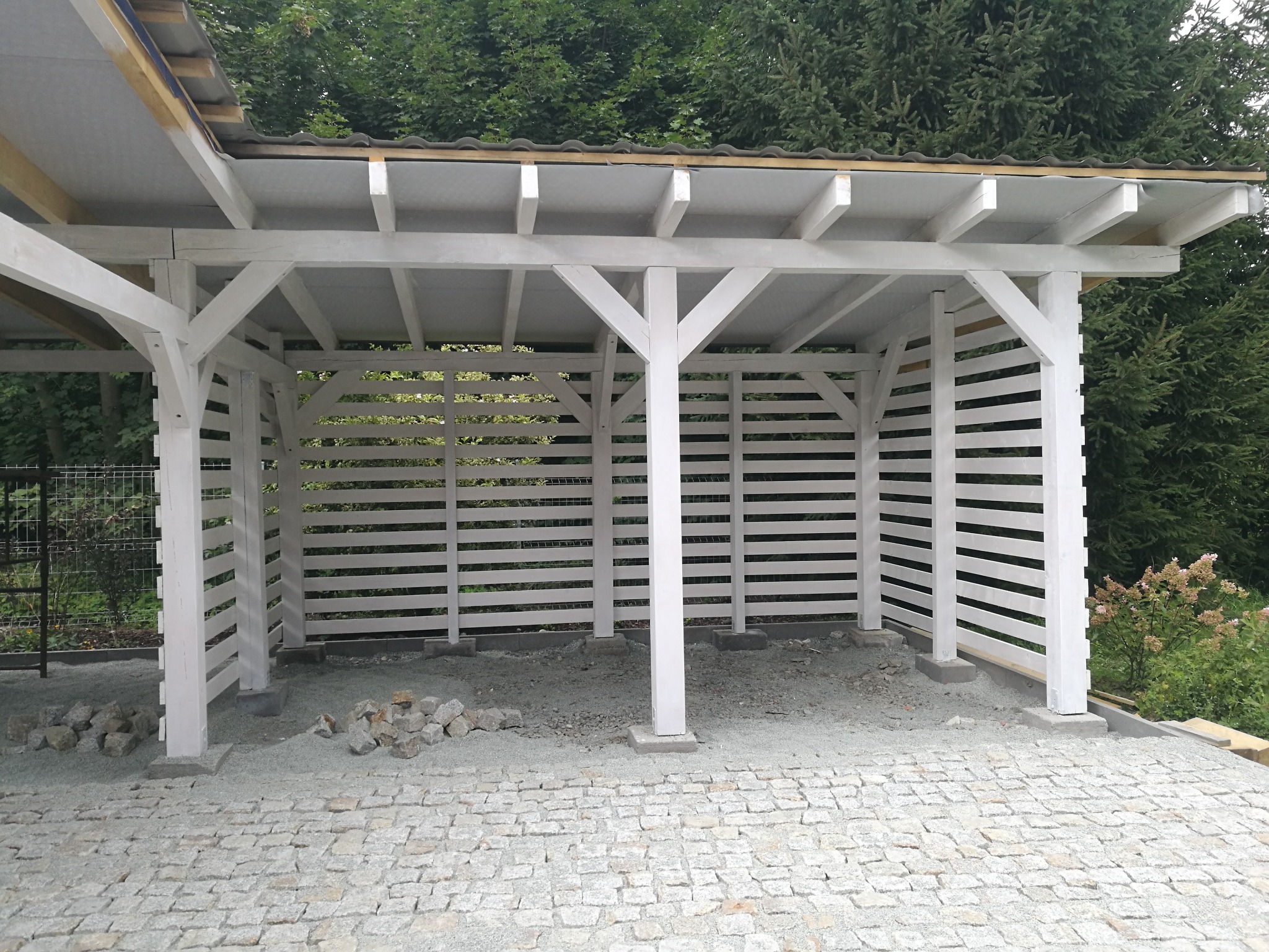 Biała, drewniana wiata z poziomymi listwami i kamienną podłogą z kostki granitowej, otoczona zielenią. Elementy konstrukcyjne w jasnym odcieniu.