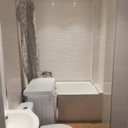 fteshhousejremonty - Niewielka łazienka z pralką obok wanny, białe płytki z delikatną falą na ścianach, drewnopodobna podłoga, zasłona prysznicowa w odcieniach szarości z motywami morskimi.