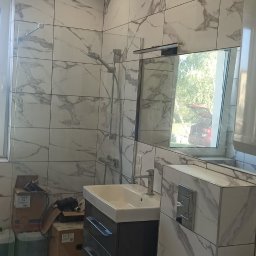 Konrad Konrad - Wykończona łazienka z marmurowymi płytkami, wisząca szafka z umywalką i lustro z widokiem na zewnątrz, kartony i folia ochronna sugerują trwające prace wykończeniowe.