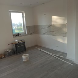 Konrad Konrad - Świeżo wykafelkowana ściana nad blatem w jasnym pomieszczeniu, widoczne białe płytki imitujące marmur, na podłodze panele imitujące drewno, przygotowane listwy przypodłogowe, wiadro z farbą...