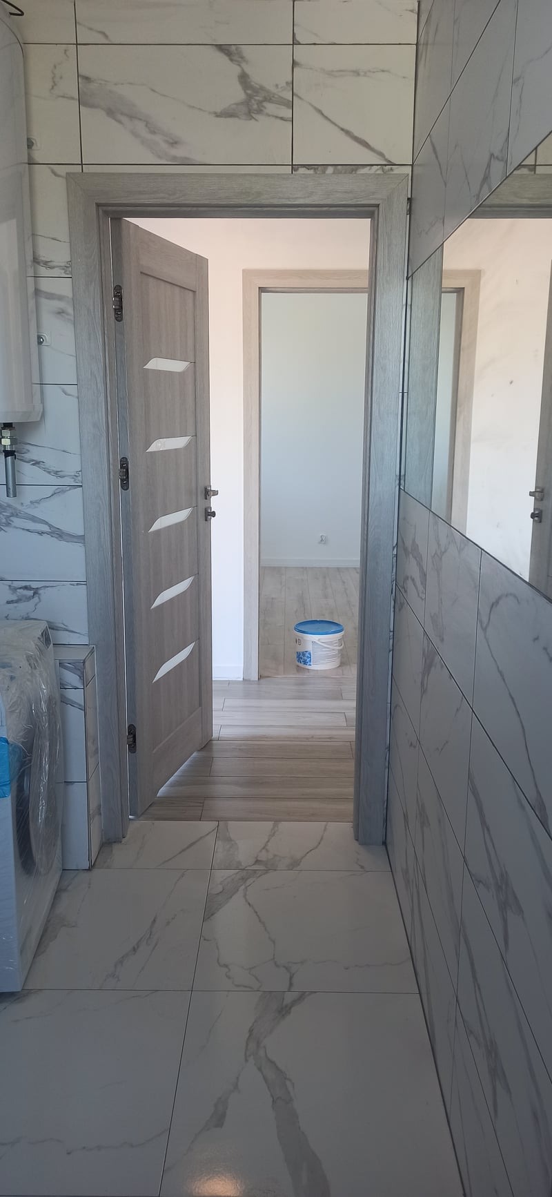 Wykończona łazienka z otwartymi drzwiami wewnętrznymi, widoczne białe marmurowe płytki na podłodze i ścianach, szara ościeżnica i drzwi z przeszkleniami oraz wiadro farby w tle.