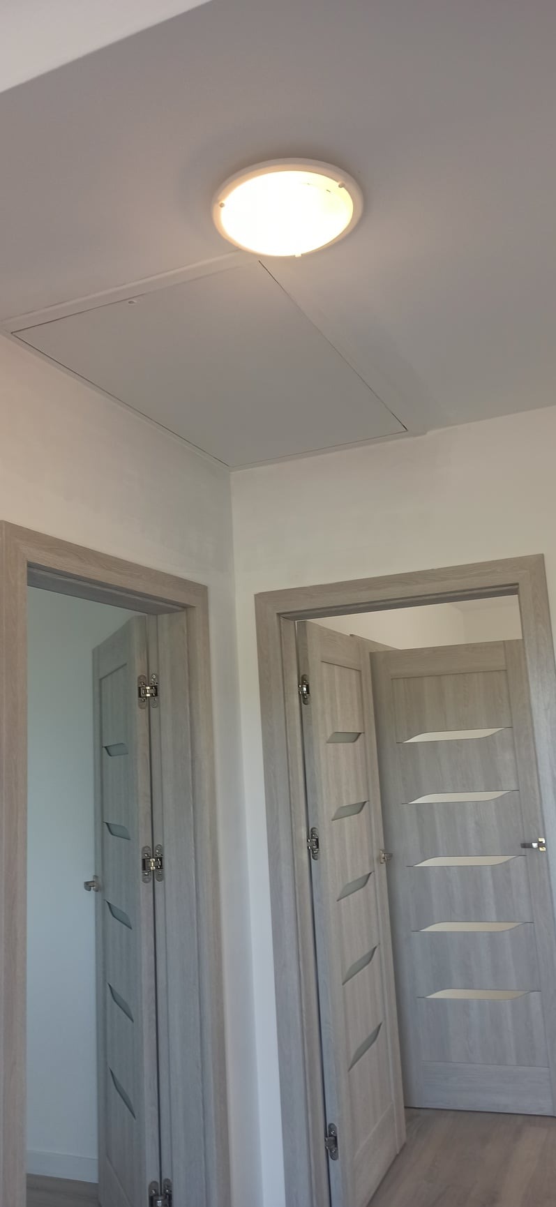 Dwa skrzydła drzwiowe z szarymi ościeżnicami i dekoracyjnymi wstawkami, jedno skrzydło otwarte, widoczna sufitowa lampa LED i klapa rewizyjna na suficie.