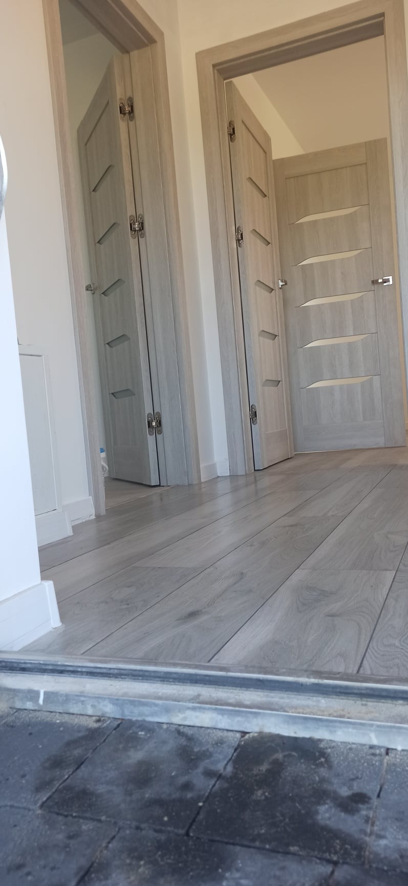 Dwa wewnętrzne drzwi z szarego drewna z poziomymi, geometrycznymi wstawkami, widoczne z niskiego kąta, z podłogą z paneli w tym samym odcieniu.