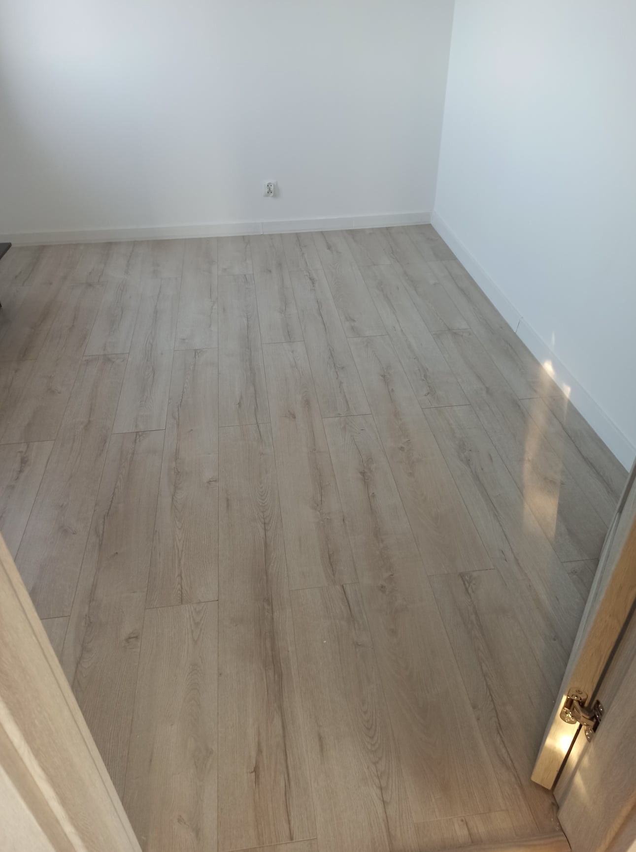 Jasne panele podłogowe ułożone w pustym pokoju z białymi ścianami i listwami przypodłogowymi, widoczne gniazdko elektryczne.