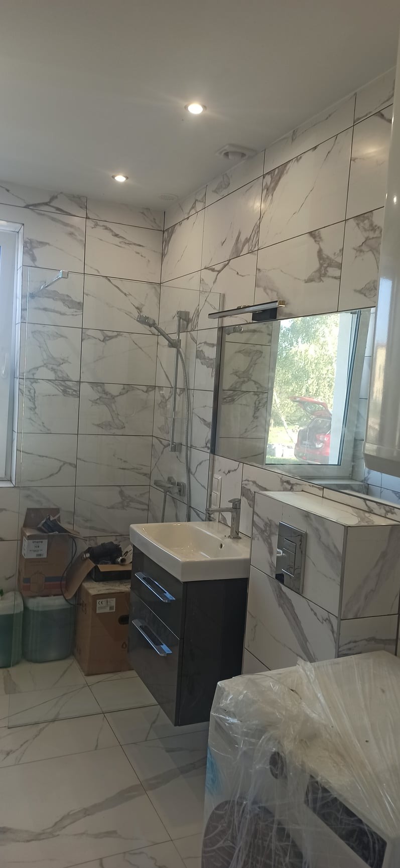 Wykończona łazienka z marmurowymi płytkami, wisząca szafka z umywalką i lustro z widokiem na zewnątrz, kartony i folia ochronna sugerują trwające prace wykończeniowe.