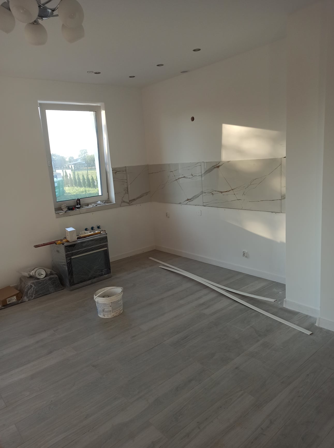 Świeżo wykafelkowana ściana nad blatem w jasnym pomieszczeniu, widoczne białe płytki imitujące marmur, na podłodze panele imitujące drewno, przygotowane listwy przypodłogowe, wiadro z farbą...