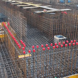 VibeTom - Skomplikowane zbrojenie fundamentów z czerwonymi osłonami na prętach, widoczne szalunki i pracownik w odblaskowej kurtce w tle. Detal budowy.